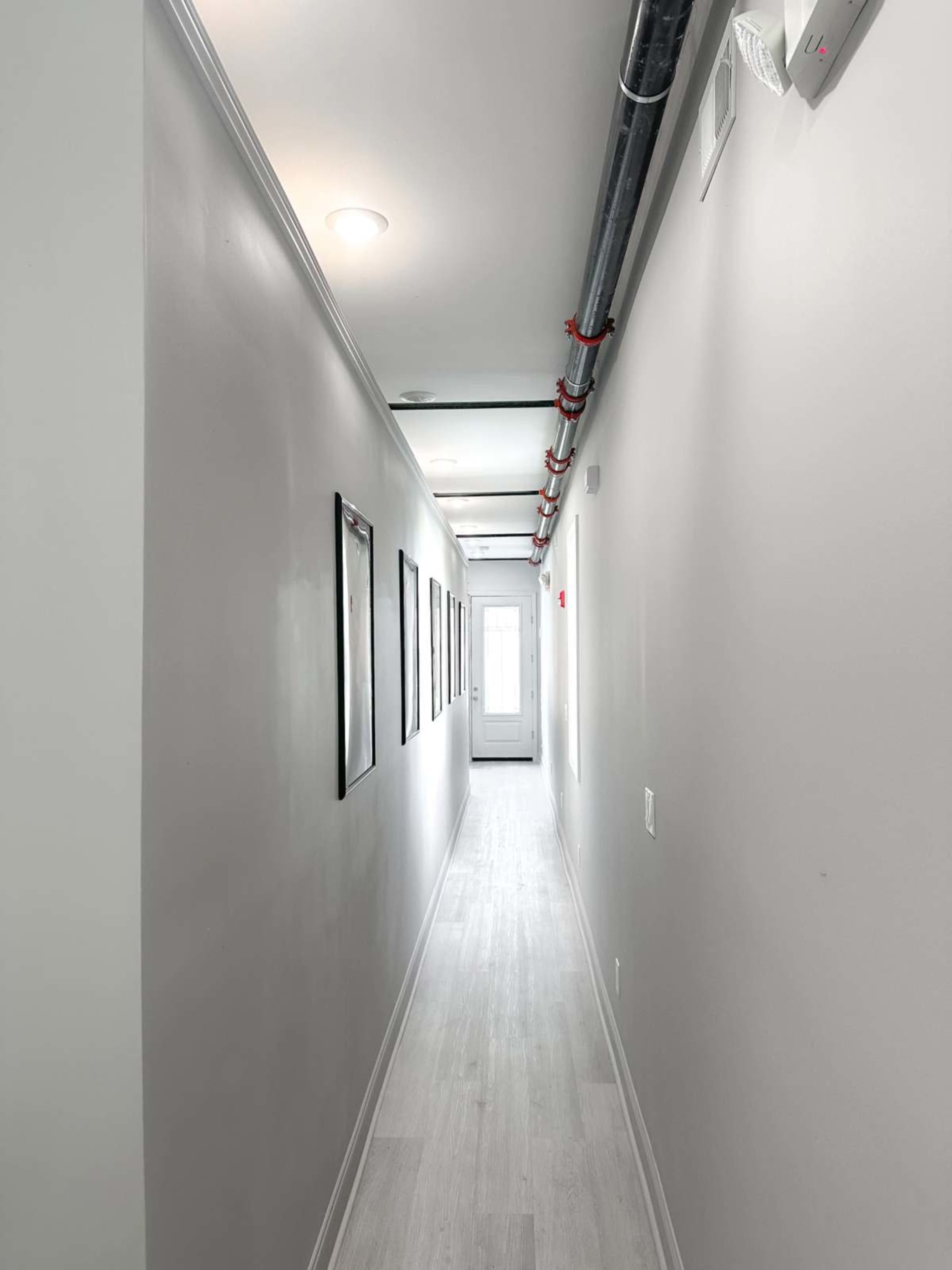 The image shows a long, narrow hallway with light gray walls, framed pictures along one side, and a door at the end.