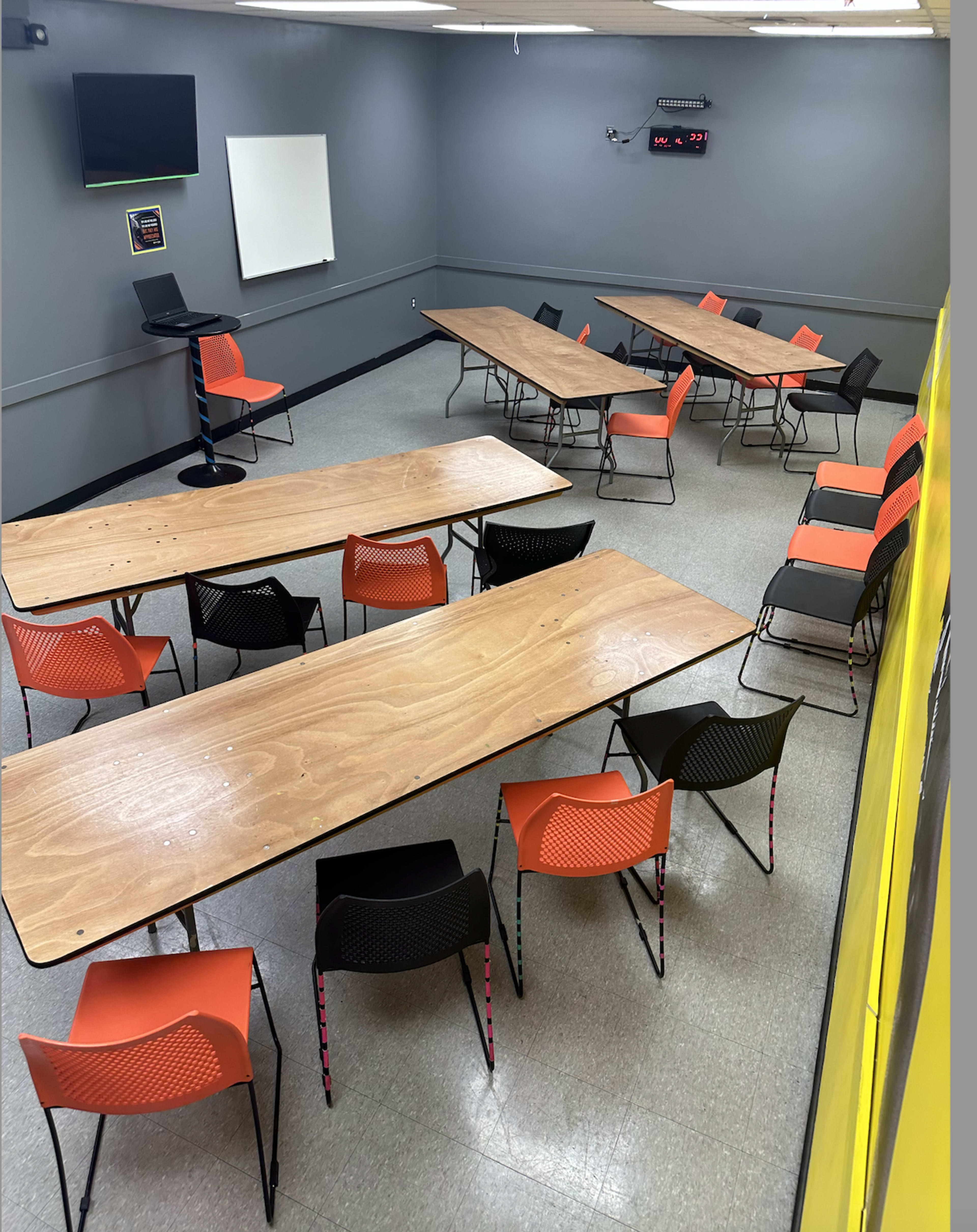 A classroom with several tables arranged in a U-shape, surrounded by chairs with orange and black colors, and equipped with a whiteboard and a screen.