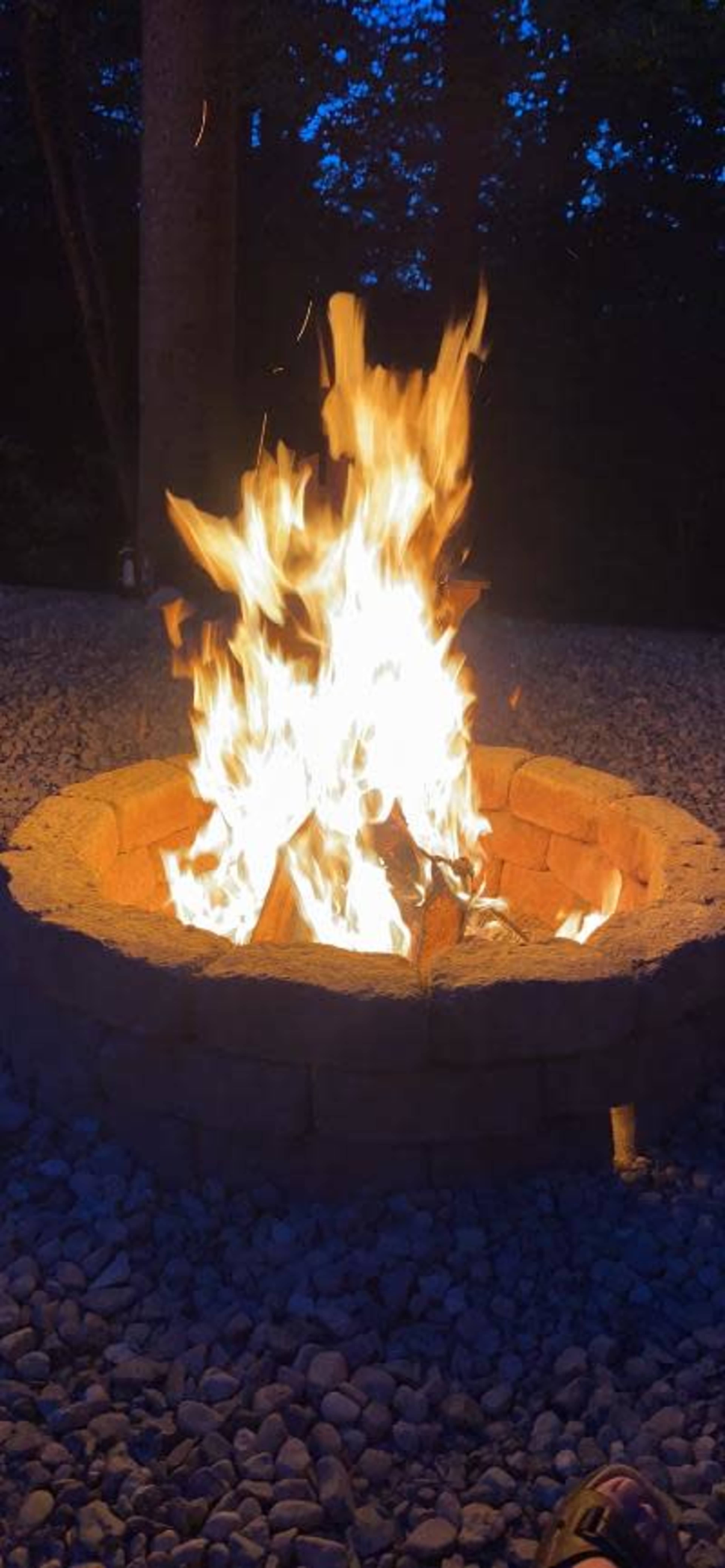 A vibrant fire burns in a circular stone fire pit surrounded by gravel in a dusky outdoor setting.