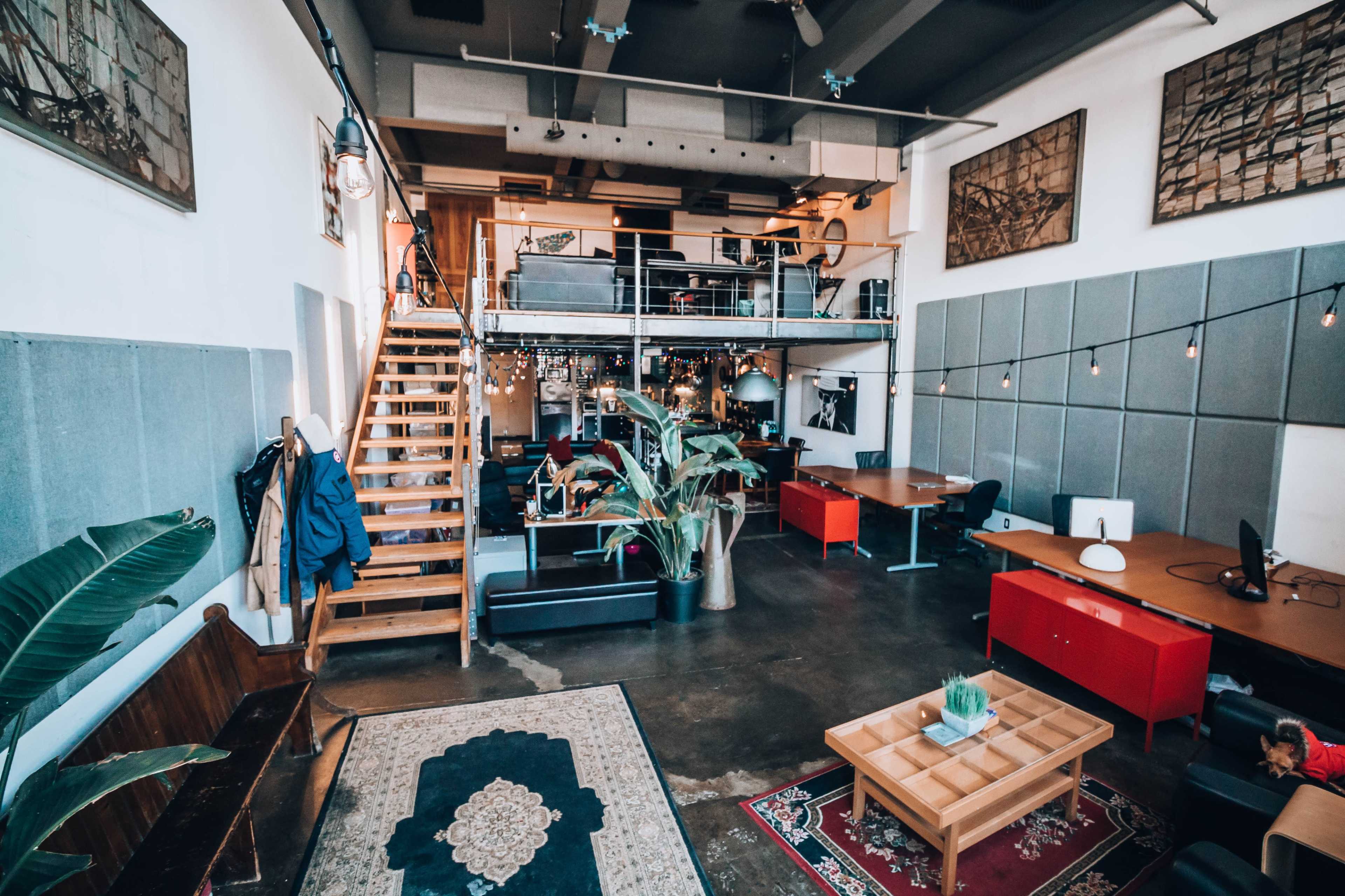 An open-plan office space with wooden stairs, multiple workstations, and decorative elements such as plants and artwork on the walls.