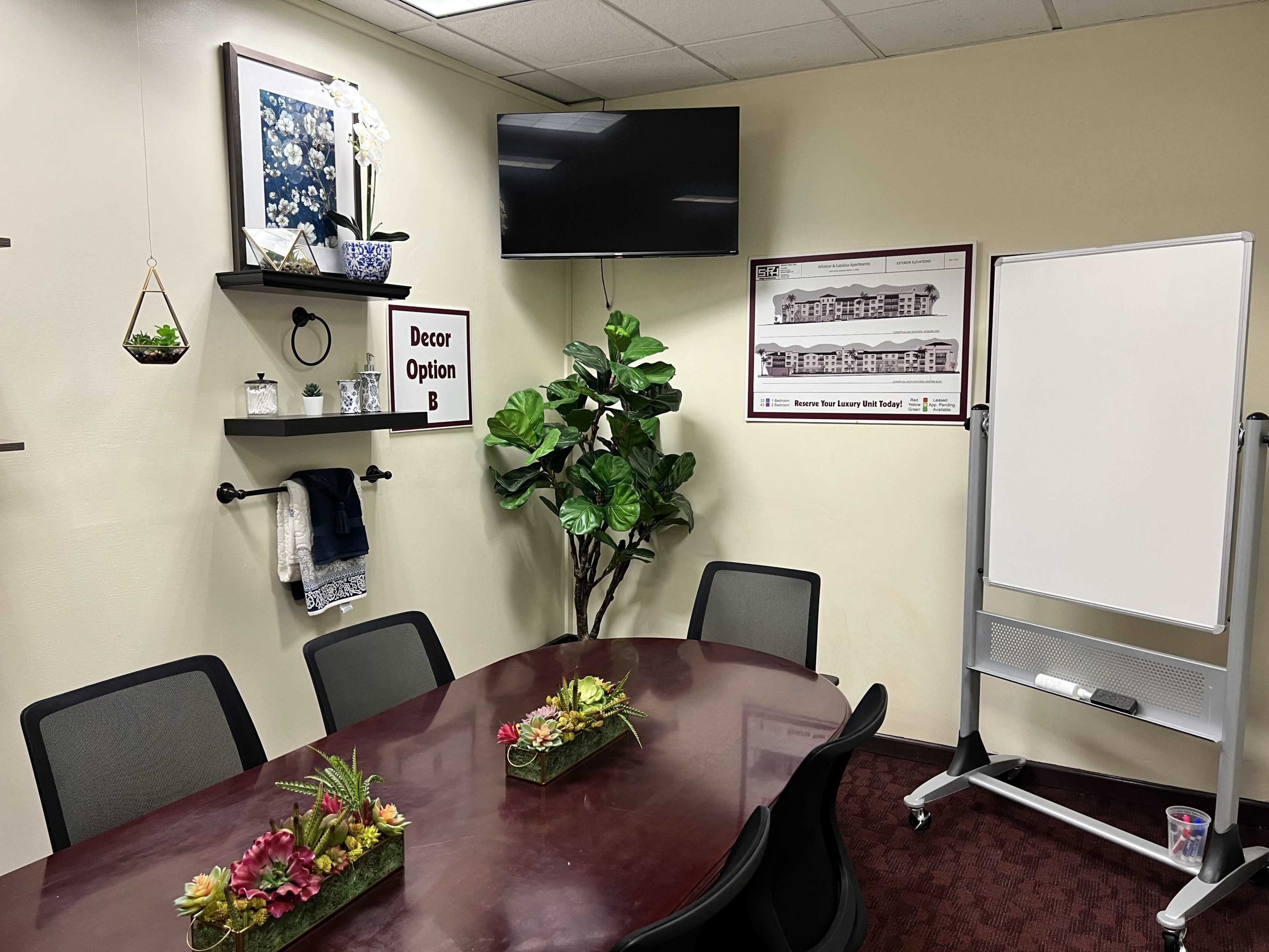 The image shows a conference room with a large table, chairs, a television on the wall, decorative plants, and whiteboard.