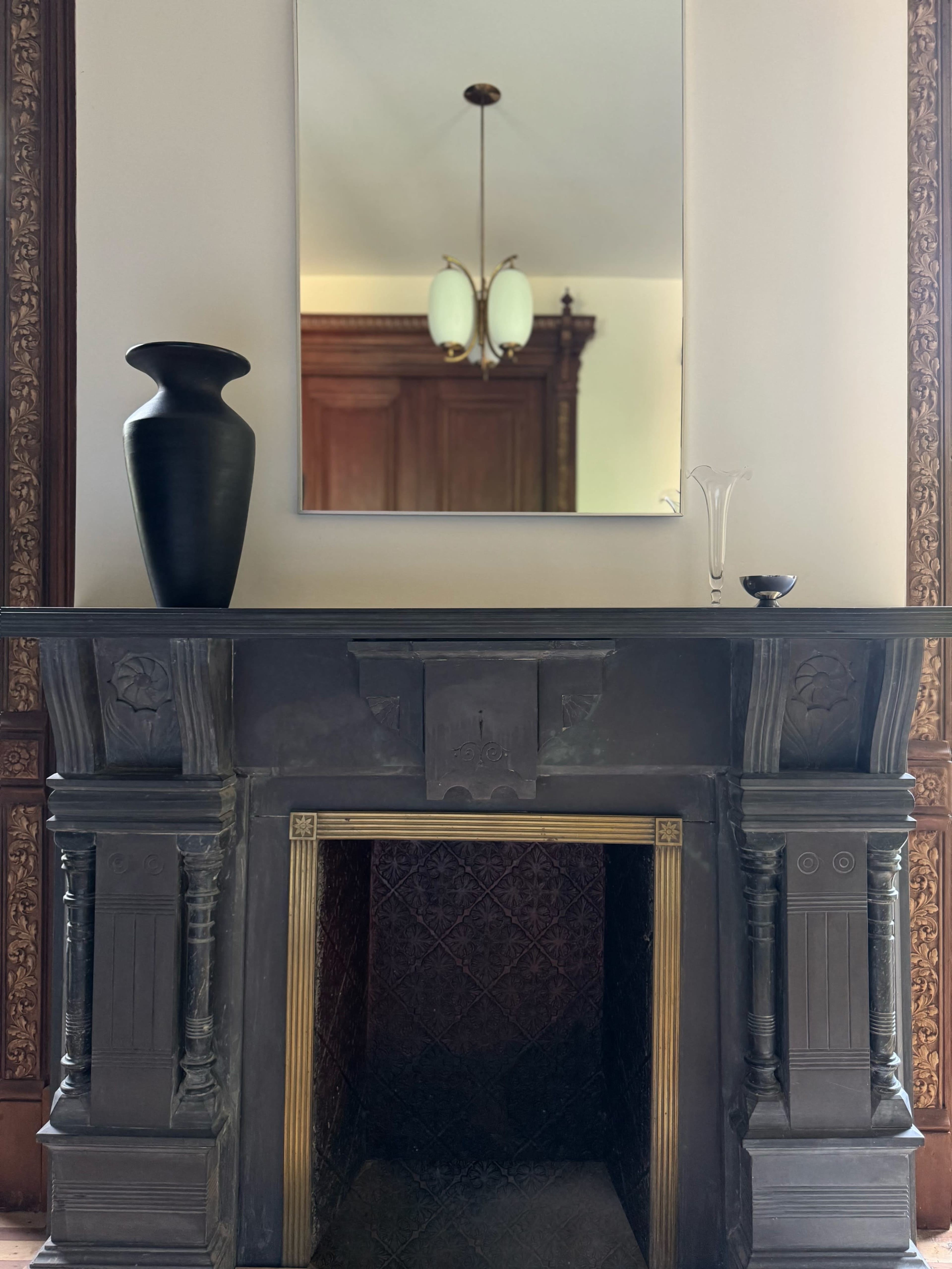 The image shows a dark stone fireplace with ornate detailing, topped by a mirror and various decorative items, including a black vase and a glass piece.