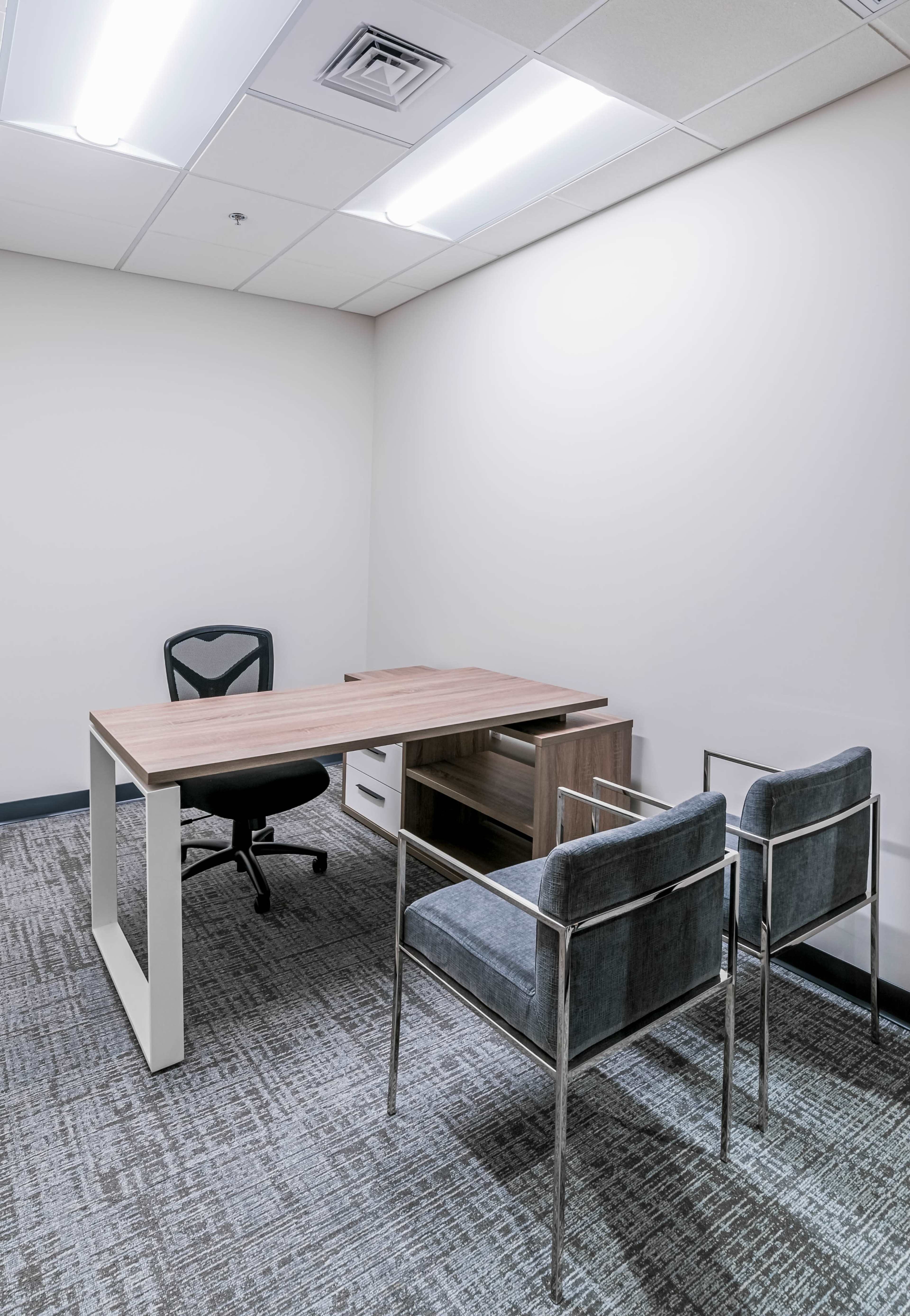 A modern office space features a wooden desk, a black office chair, and two gray upholstered chairs against a plain wall.