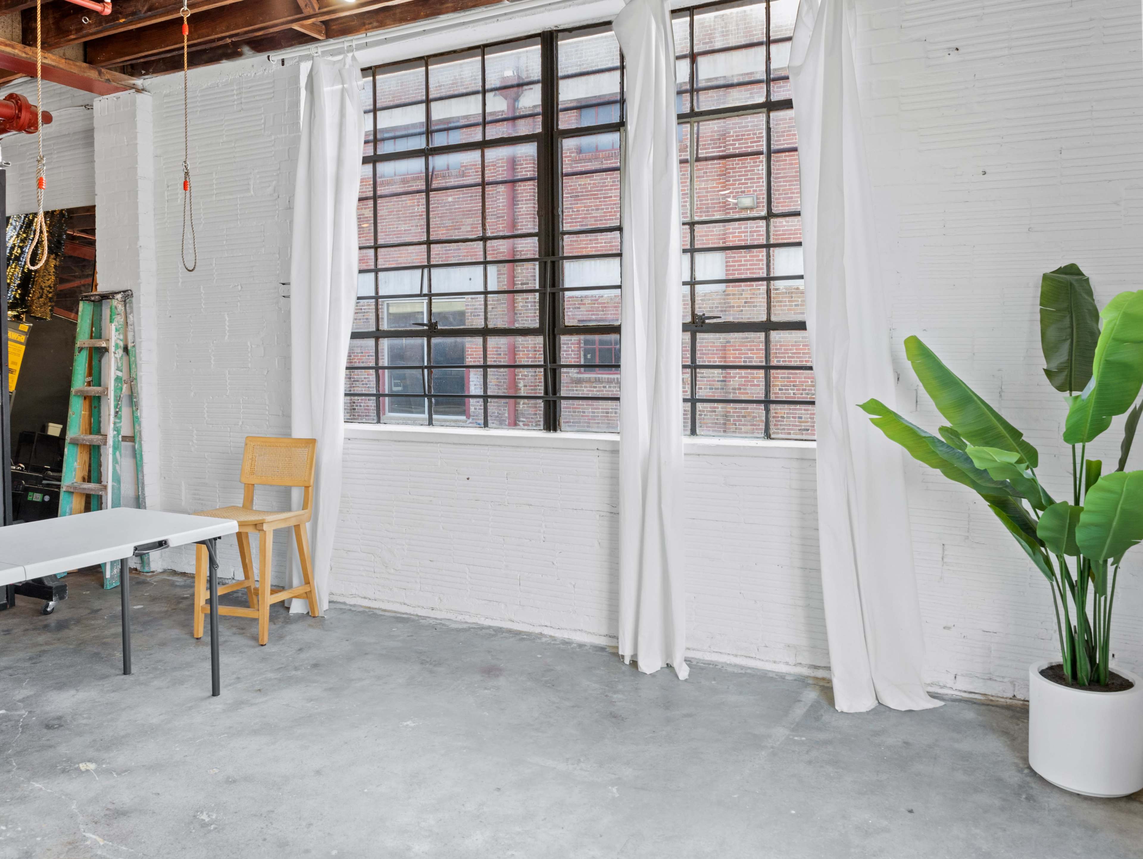A bright industrial space featuring large windows with sheer white curtains, a potted plant, a small wooden chair, and a simple table on a concrete floor.