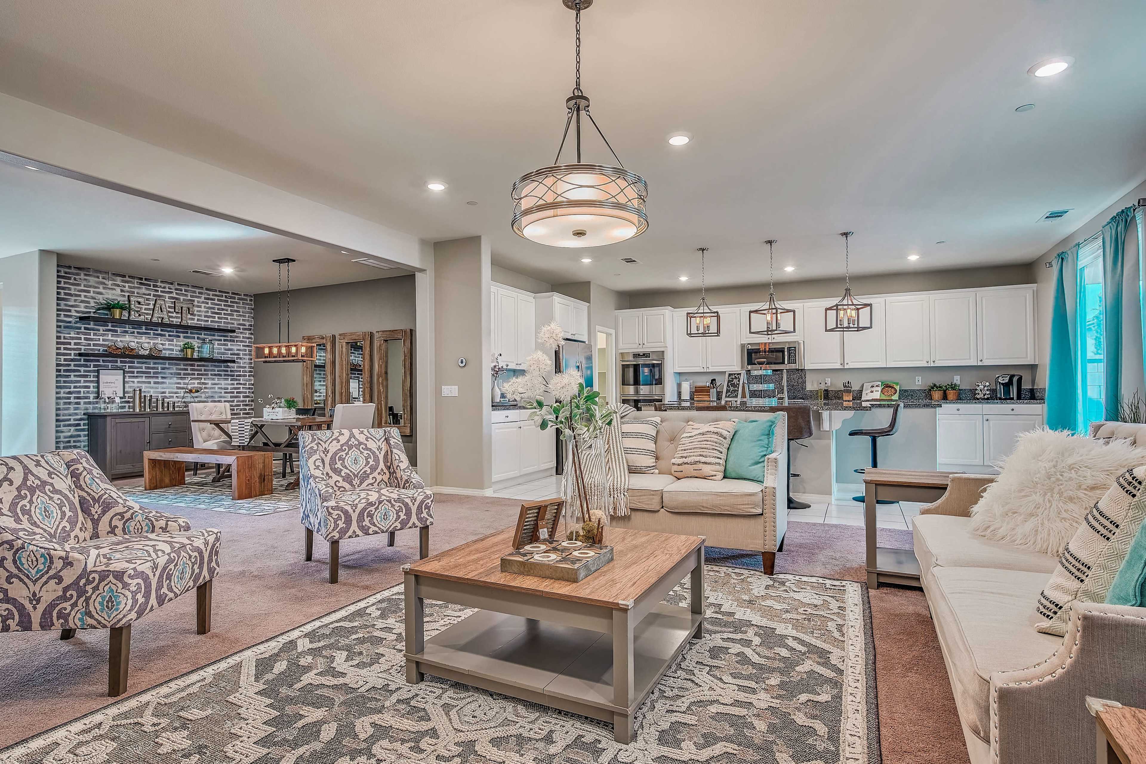 The image shows a modern living room with a dining area, featuring patterned chairs, a coffee table, and an open kitchen in the background.