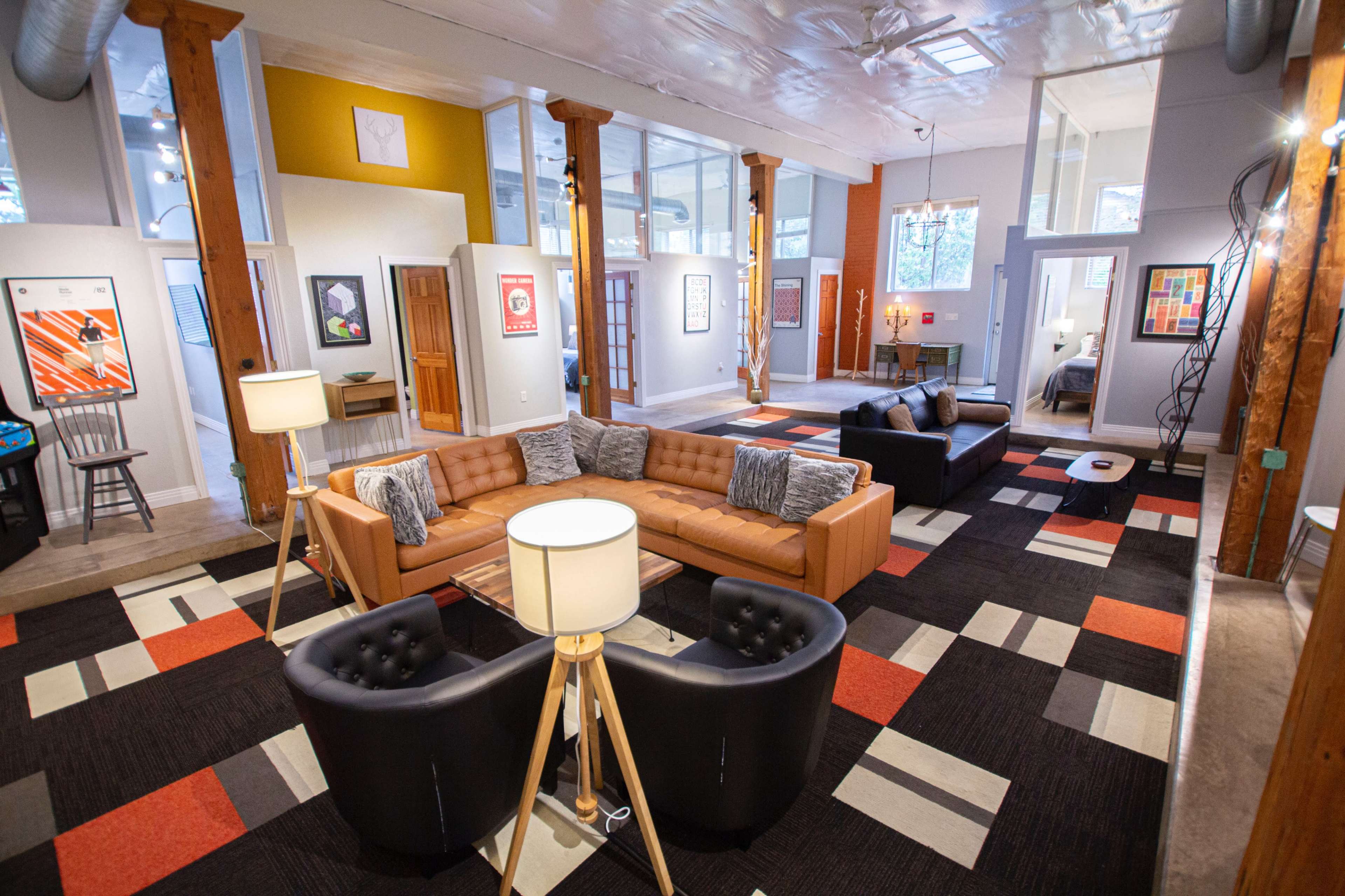 The image shows a spacious modern living room with a combination of a brown sectional sofa, black chairs, and colorful carpet, illuminated by lamps and natural light.