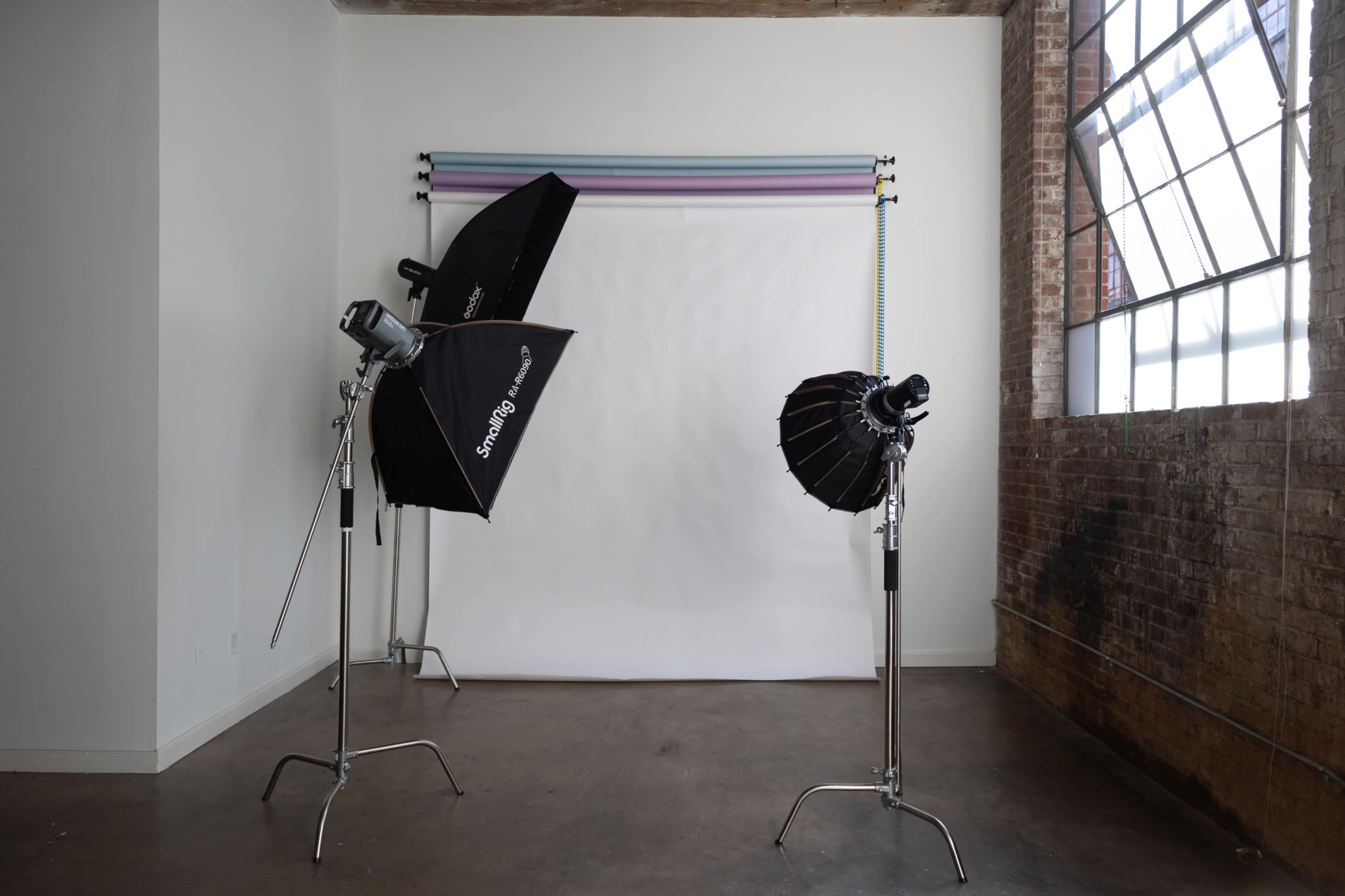 A photography studio features two lighting setups pointing at a blank backdrop with a large window providing natural light.