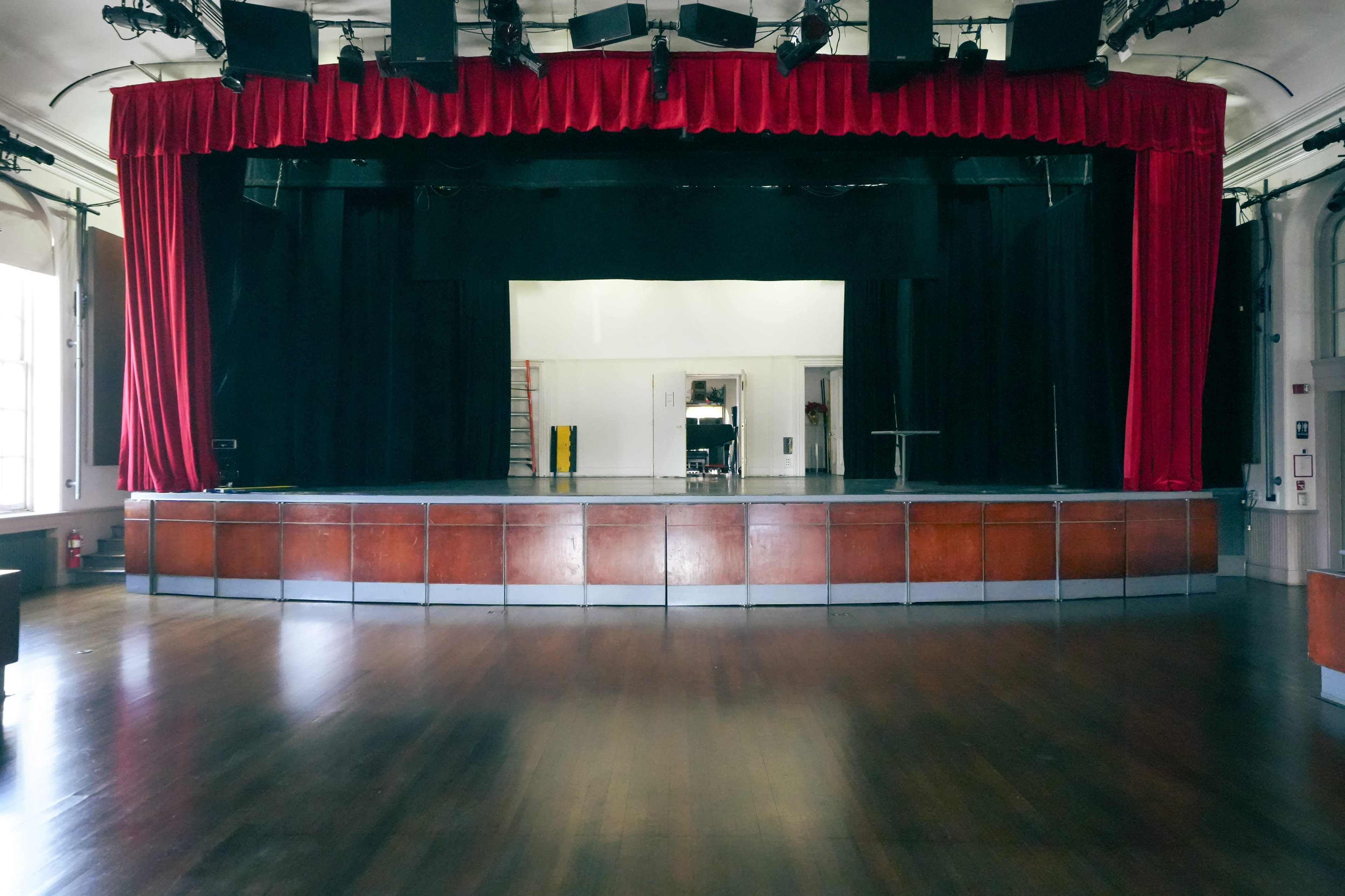 A dark theater stage with a red curtain, wooden flooring, and a backdrop that appears to be empty and undisturbed.