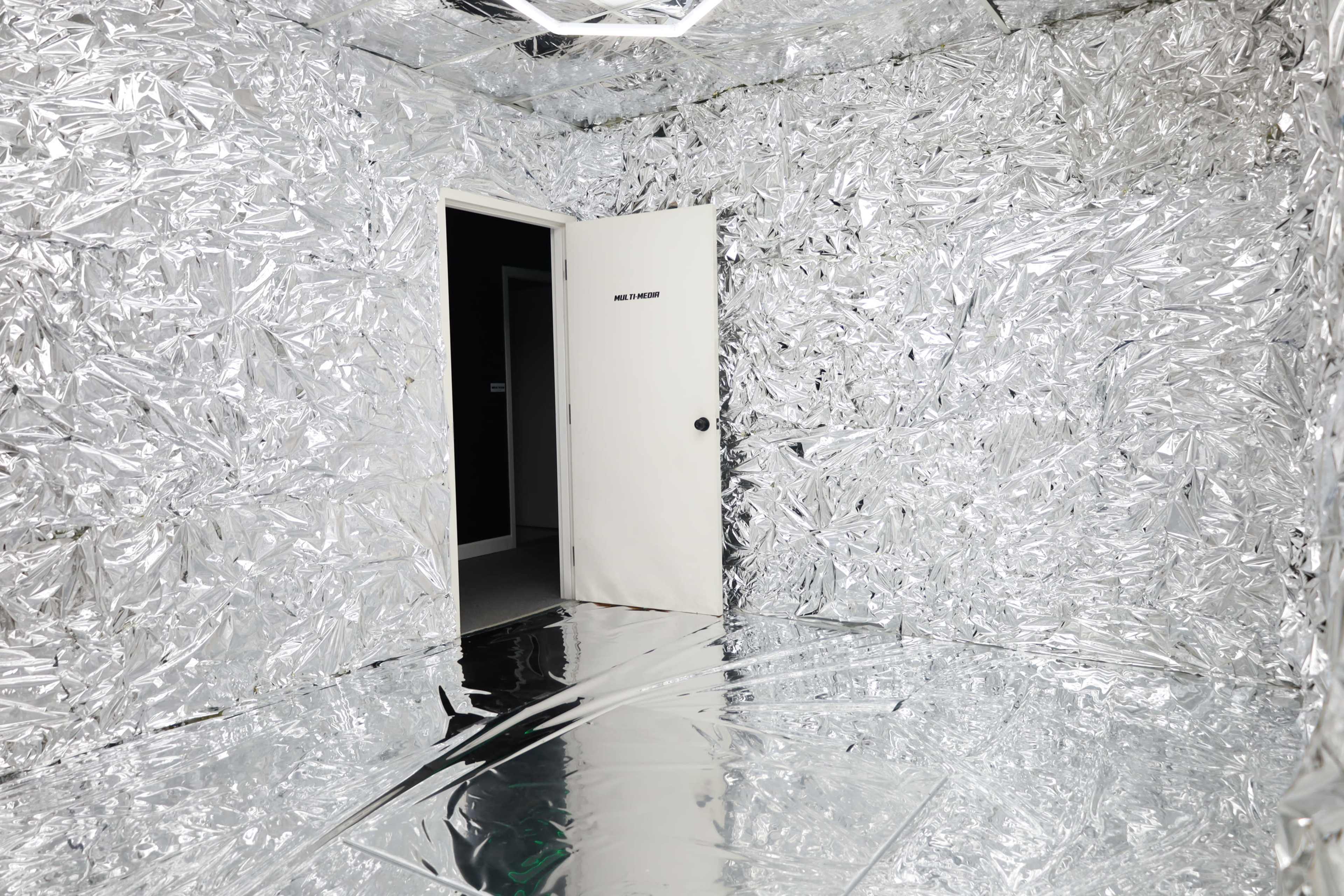 The image shows a room completely lined with crumpled silver foil, featuring a single closed door on one wall.