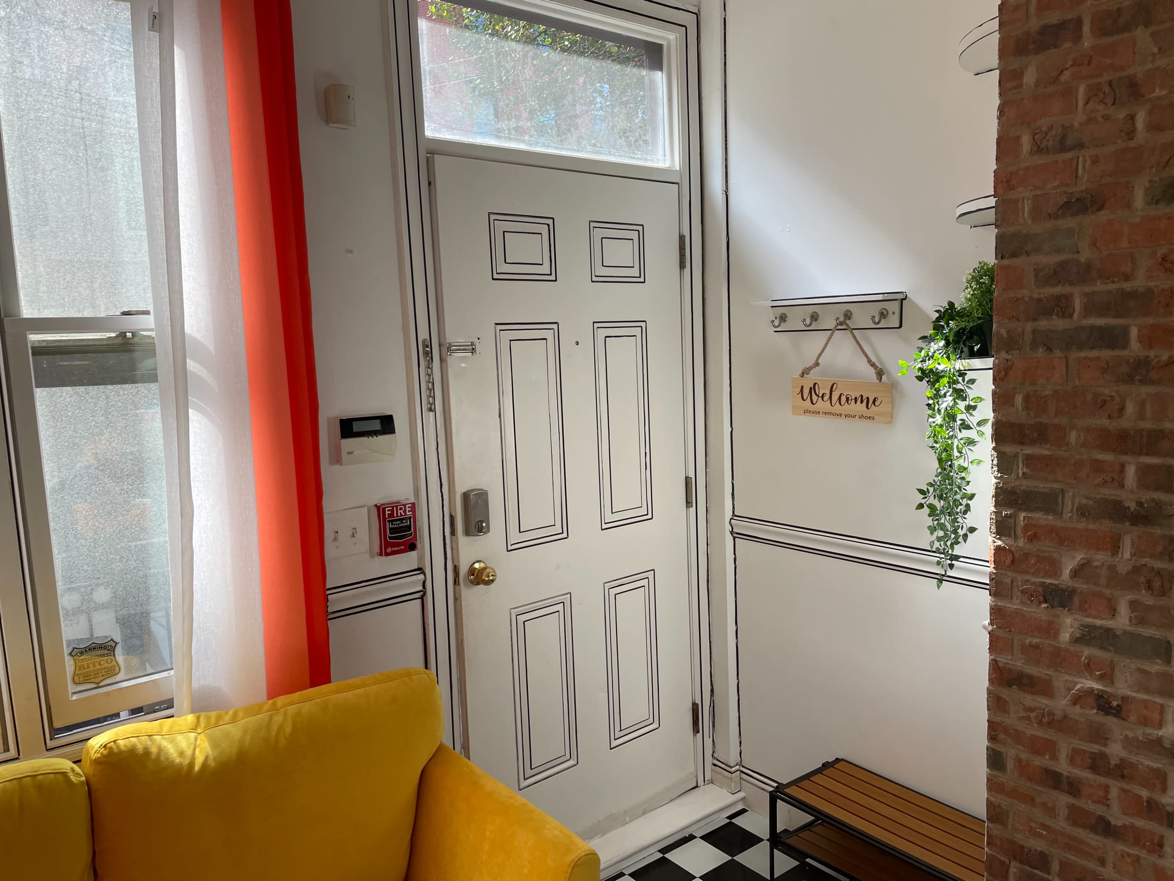 An entryway with a white door, a yellow couch, checkered floor tiles, and a small shelf with plants.