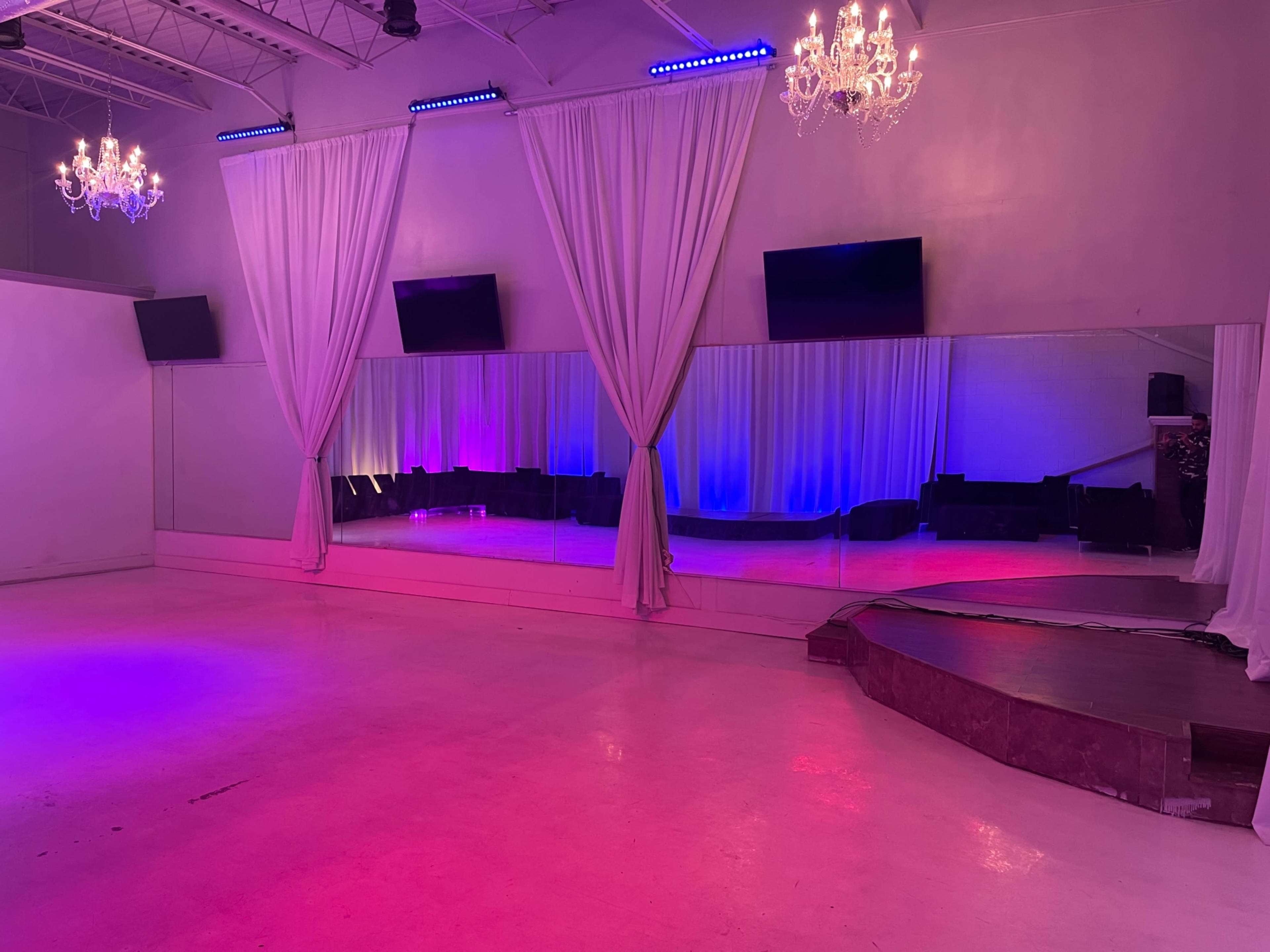 The image shows a spacious dance studio with large mirrors, purple and pink lighting, and draped curtains.