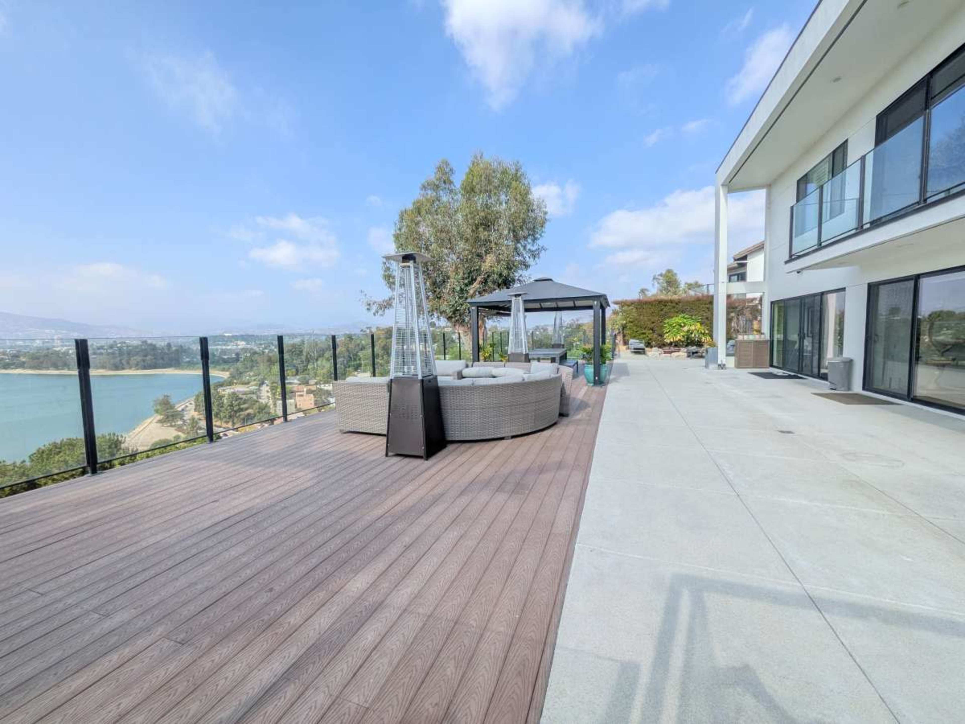 The image shows a spacious outdoor deck area with a view of a lake, featuring lounge seating, a gazebo, and large glass railings.