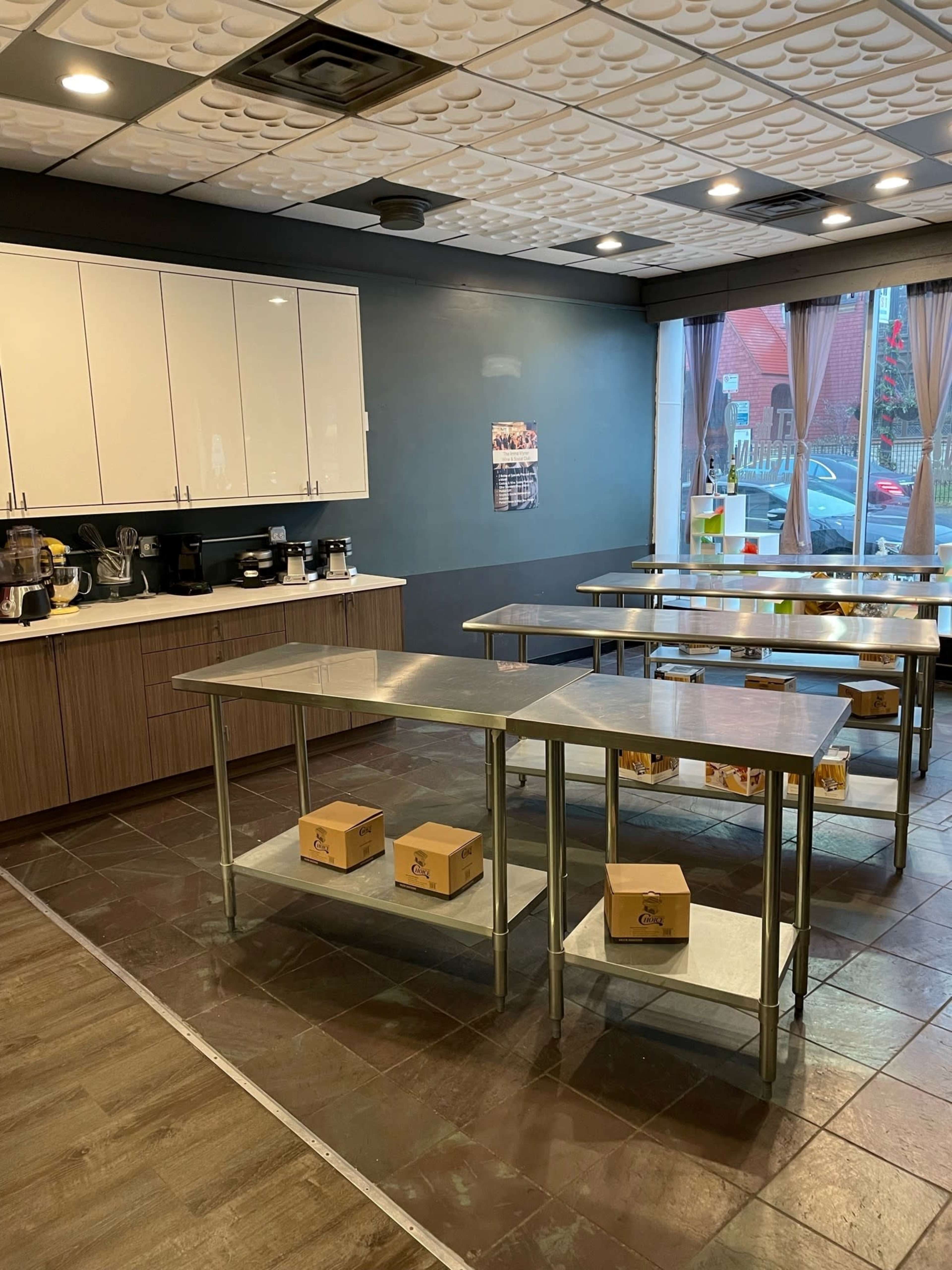 The image shows a kitchen space with stainless steel tables and several boxes arranged on the lower shelves, alongside a countertop with various kitchen appliances and cabinets.