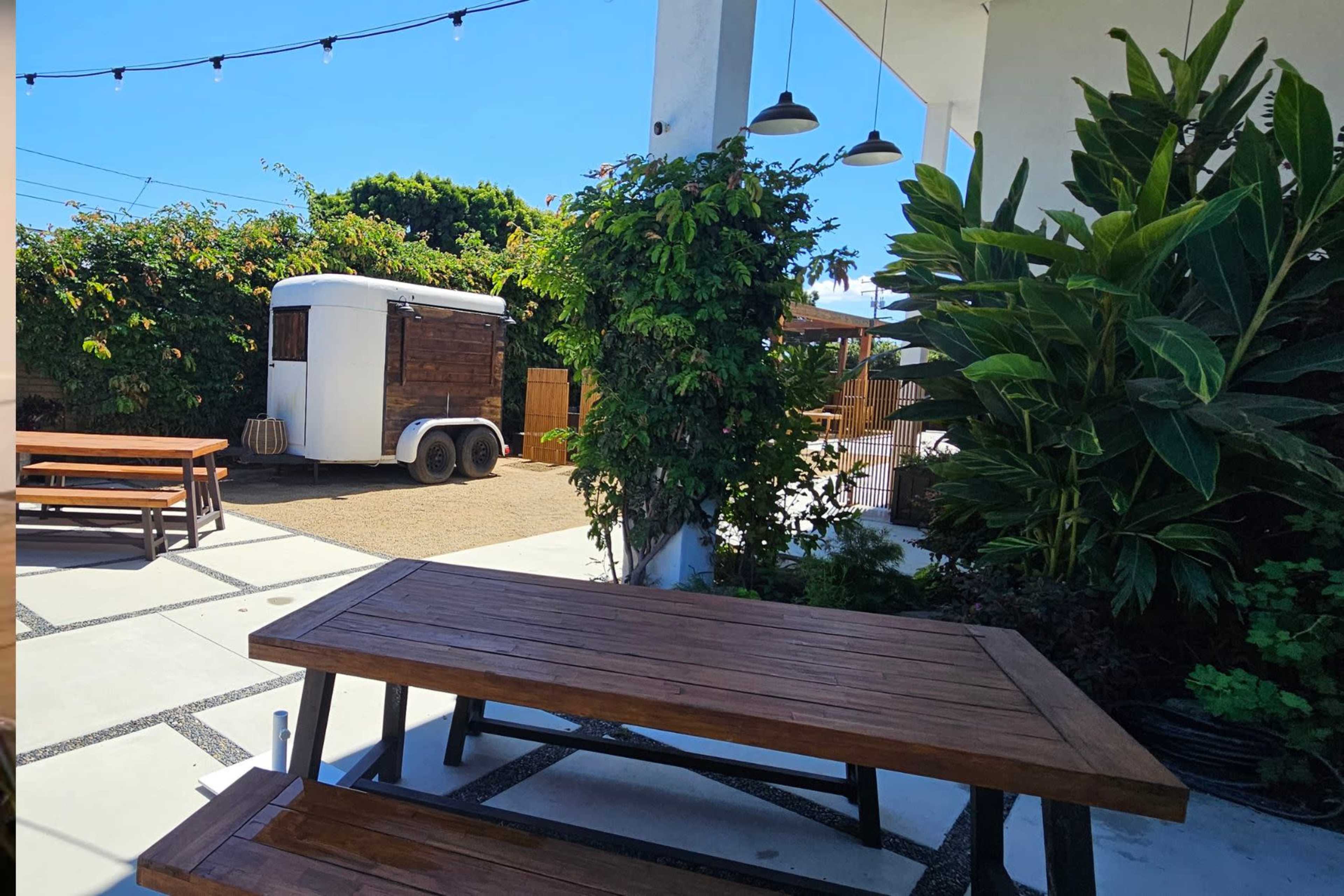 A wooden table and benches are set in a bright outdoor area featuring greenery and a trailer in the background.