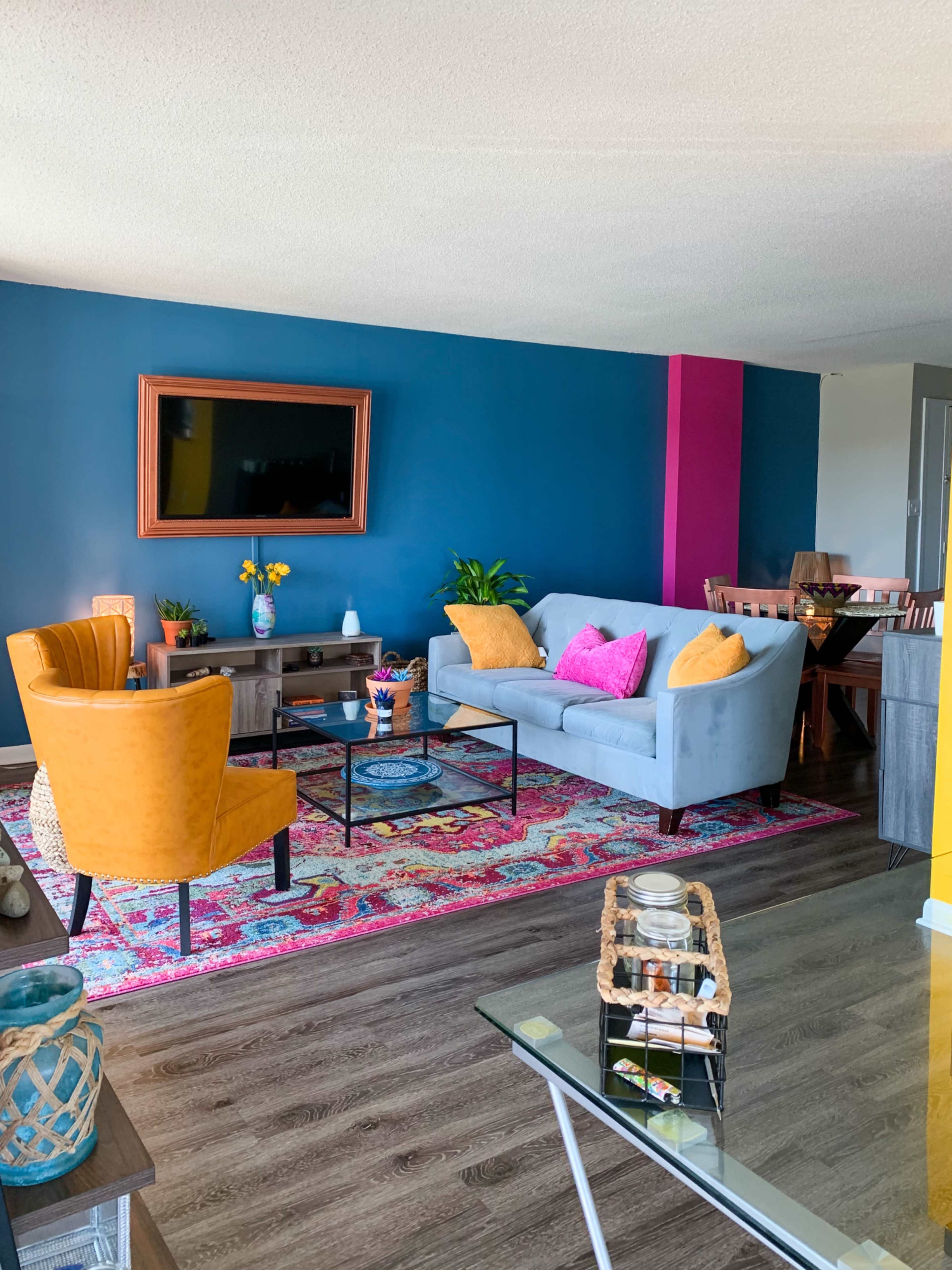 The living room features a blue accent wall, colorful decor, a gray sofa, two yellow armchairs, and a patterned area rug.