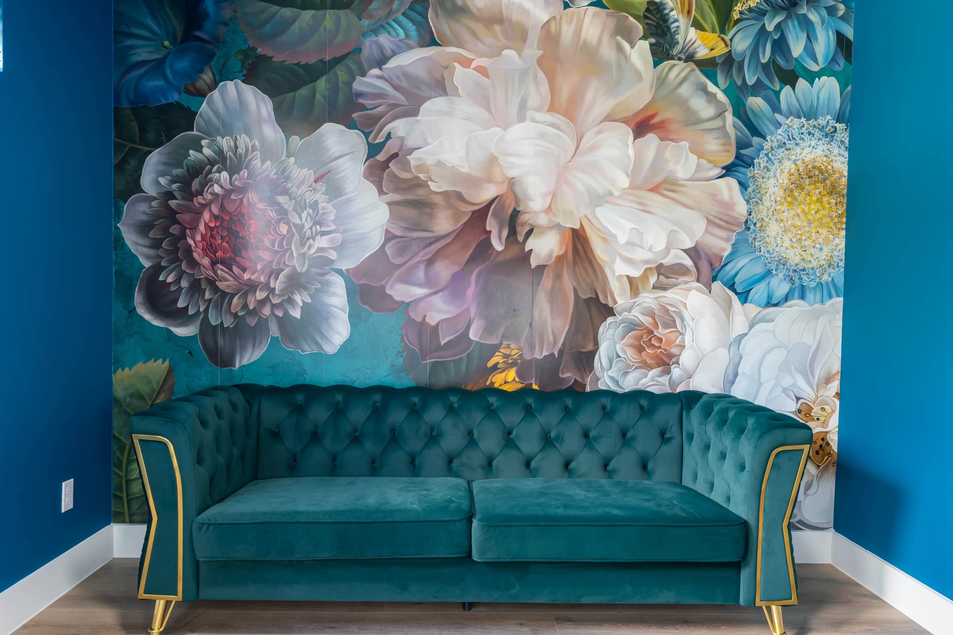 A teal velvet sofa is positioned against a wall adorned with large, vibrant floral murals.