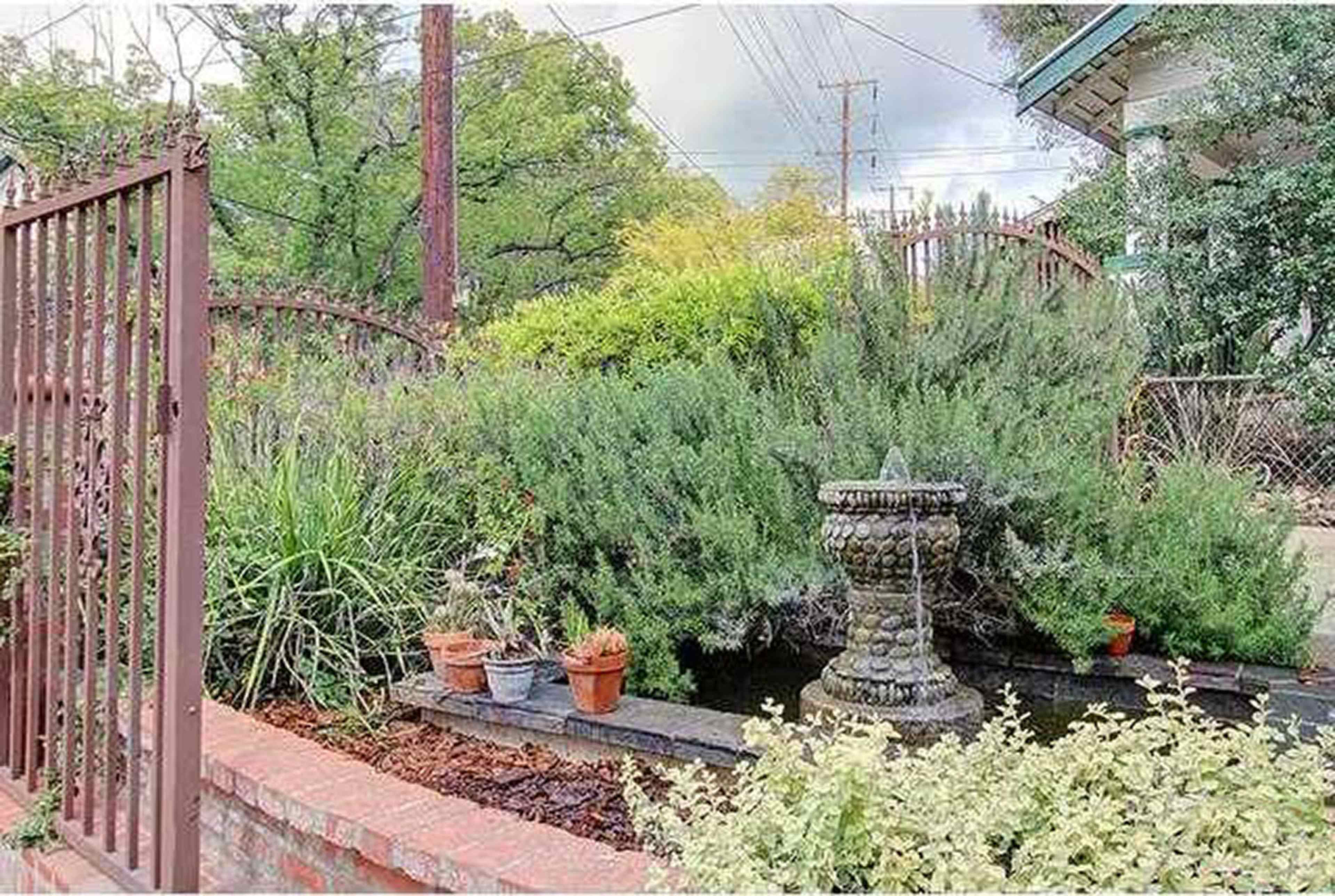 A stone fountain sits amidst lush greenery and potted plants in a garden enclosed by a wrought iron fence.