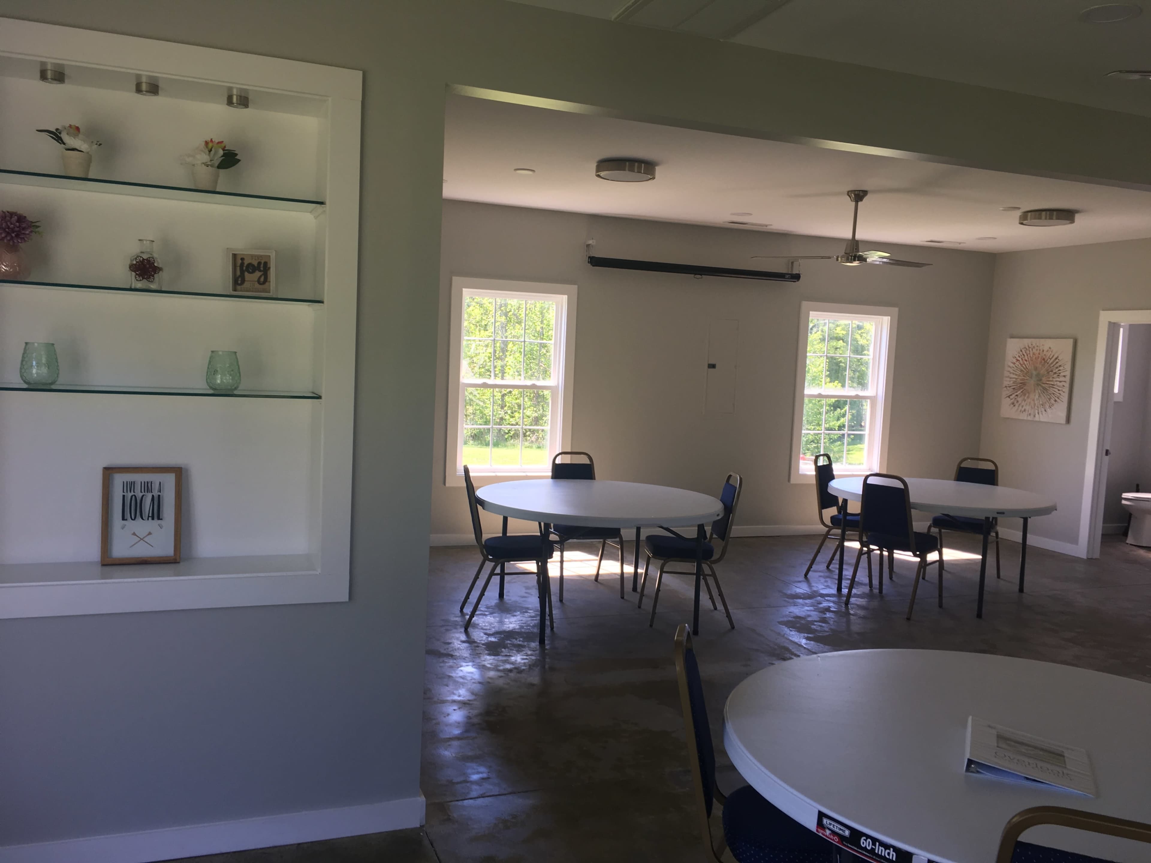 A spacious room with white walls, two round tables set up with chairs, and windows allowing natural light in.
