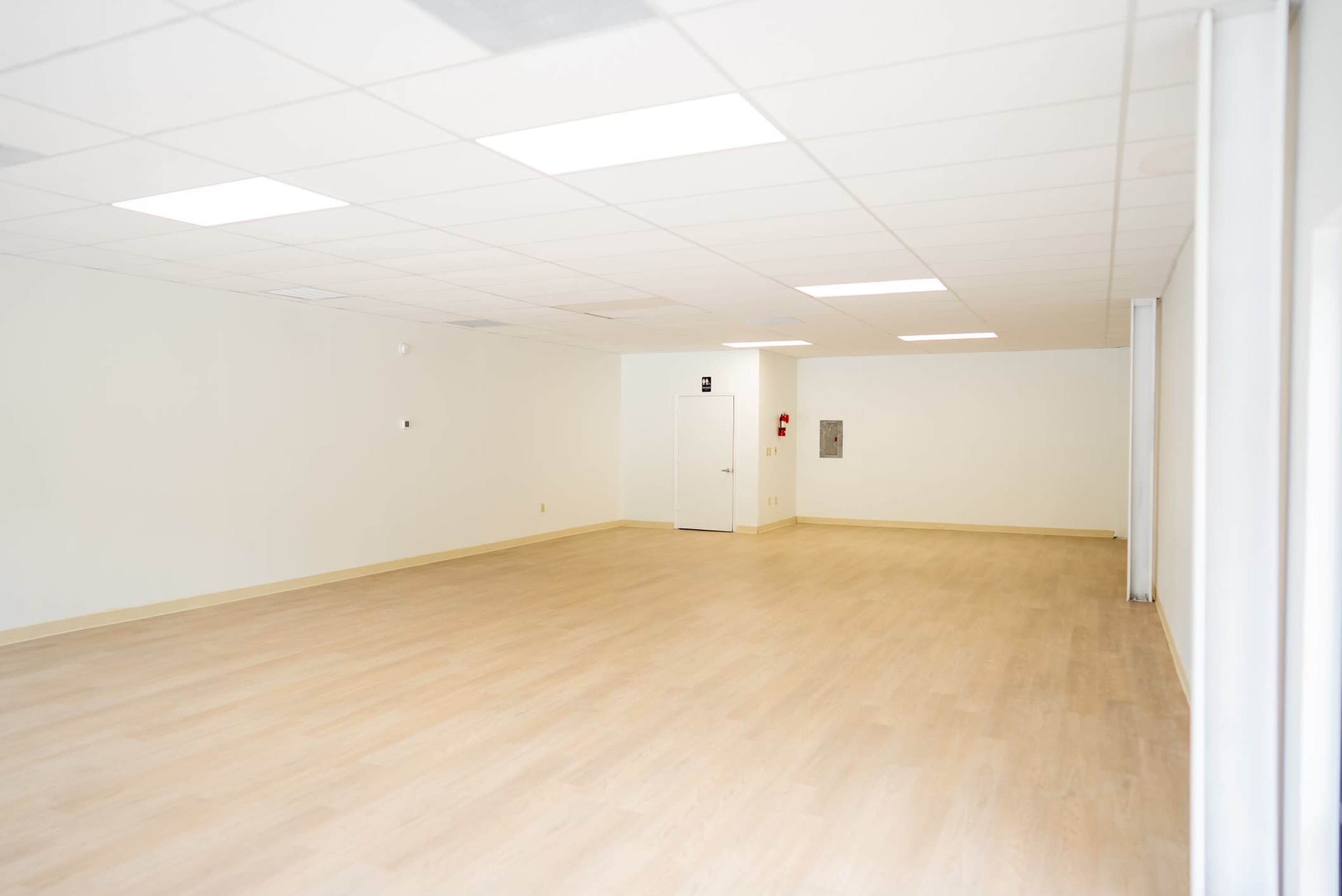 The image shows a vacant room with smooth wooden flooring, white walls, and a few light fixtures on the ceiling.