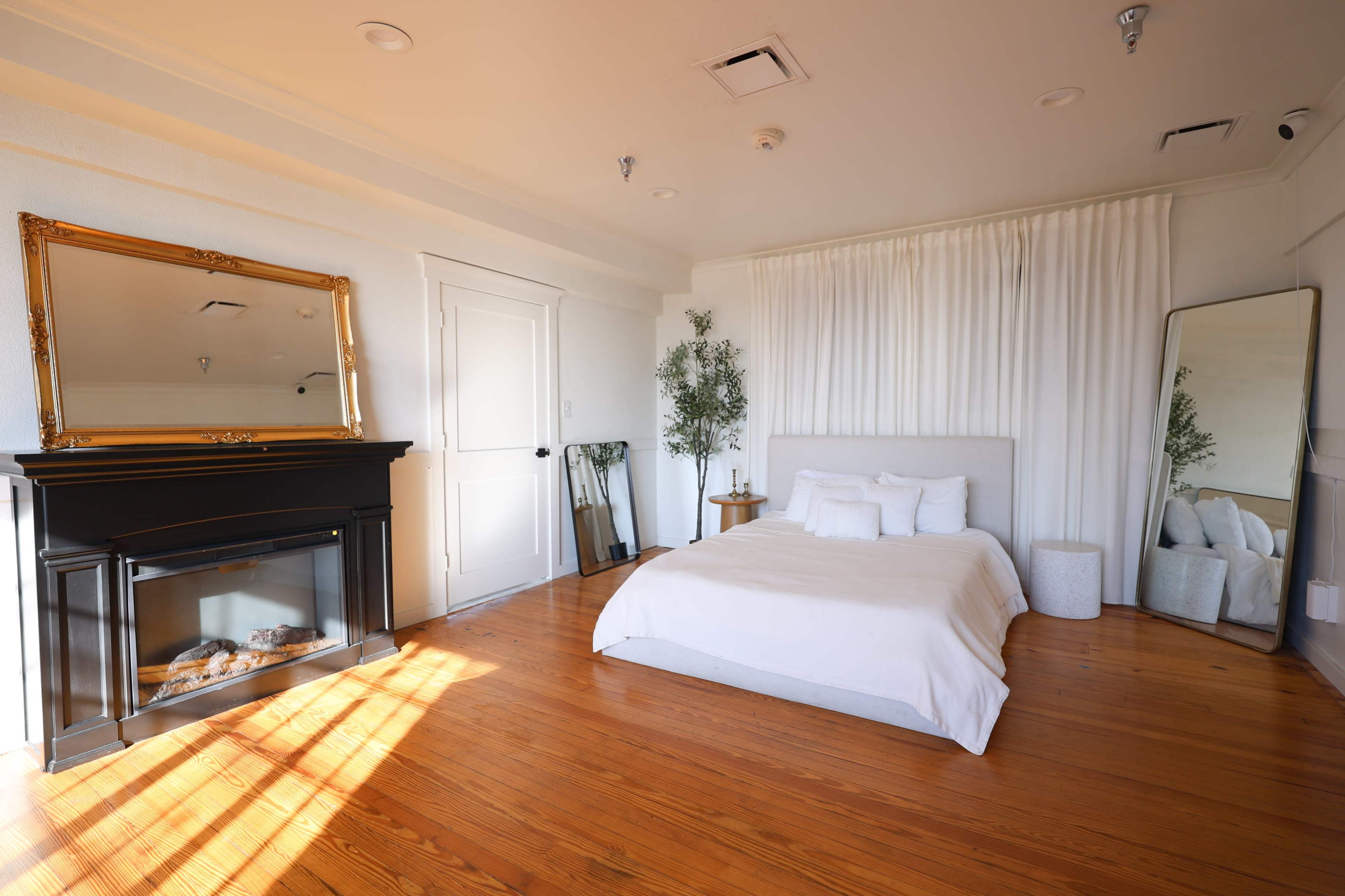 The image shows a minimalist bedroom featuring a bed with white linens, a decorative mirror, a fireplace, and wooden flooring.