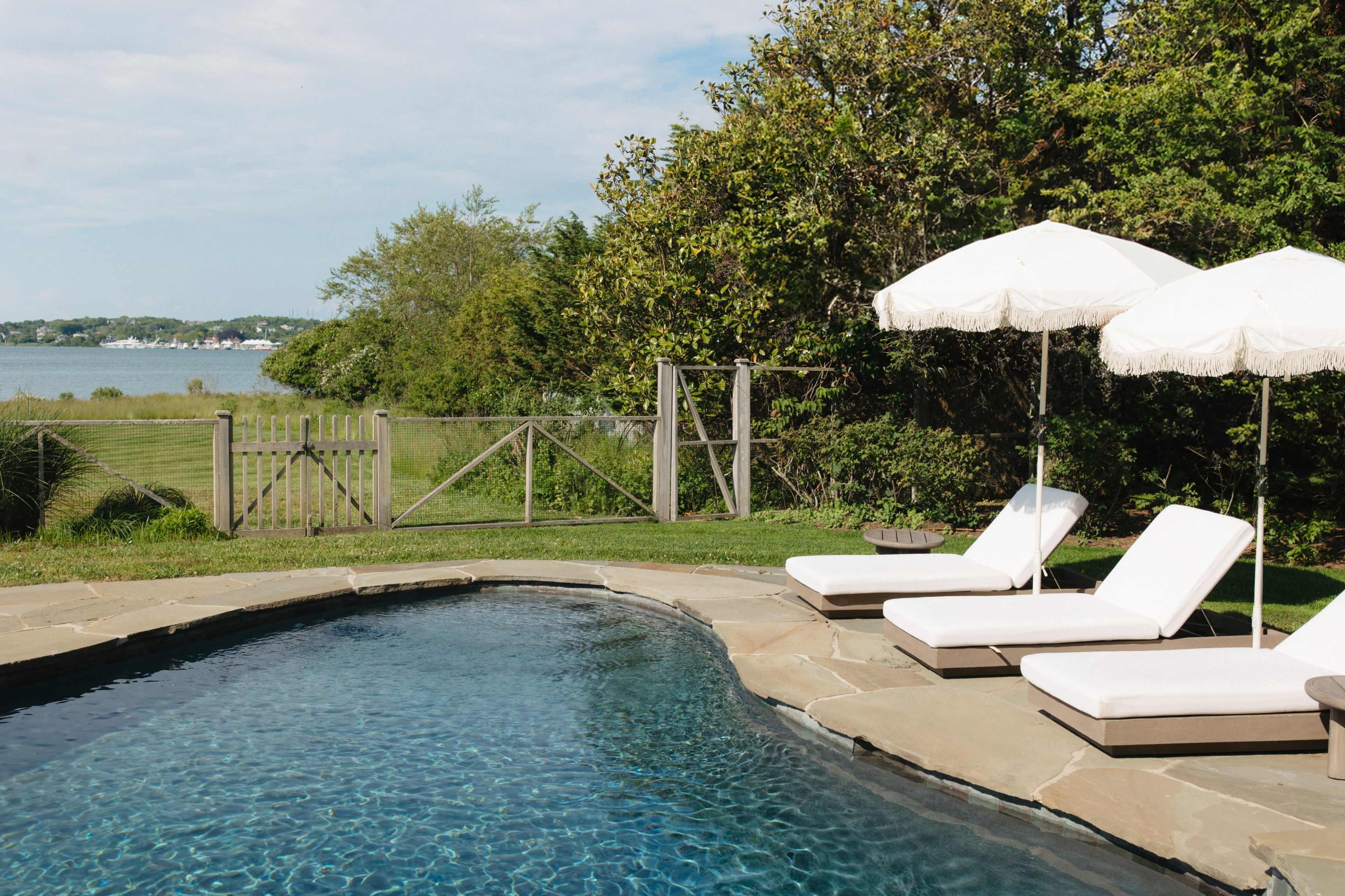 A swimming pool with two white lounge chairs and umbrellas is situated next to a grassy area overlooking a body of water and distant trees.