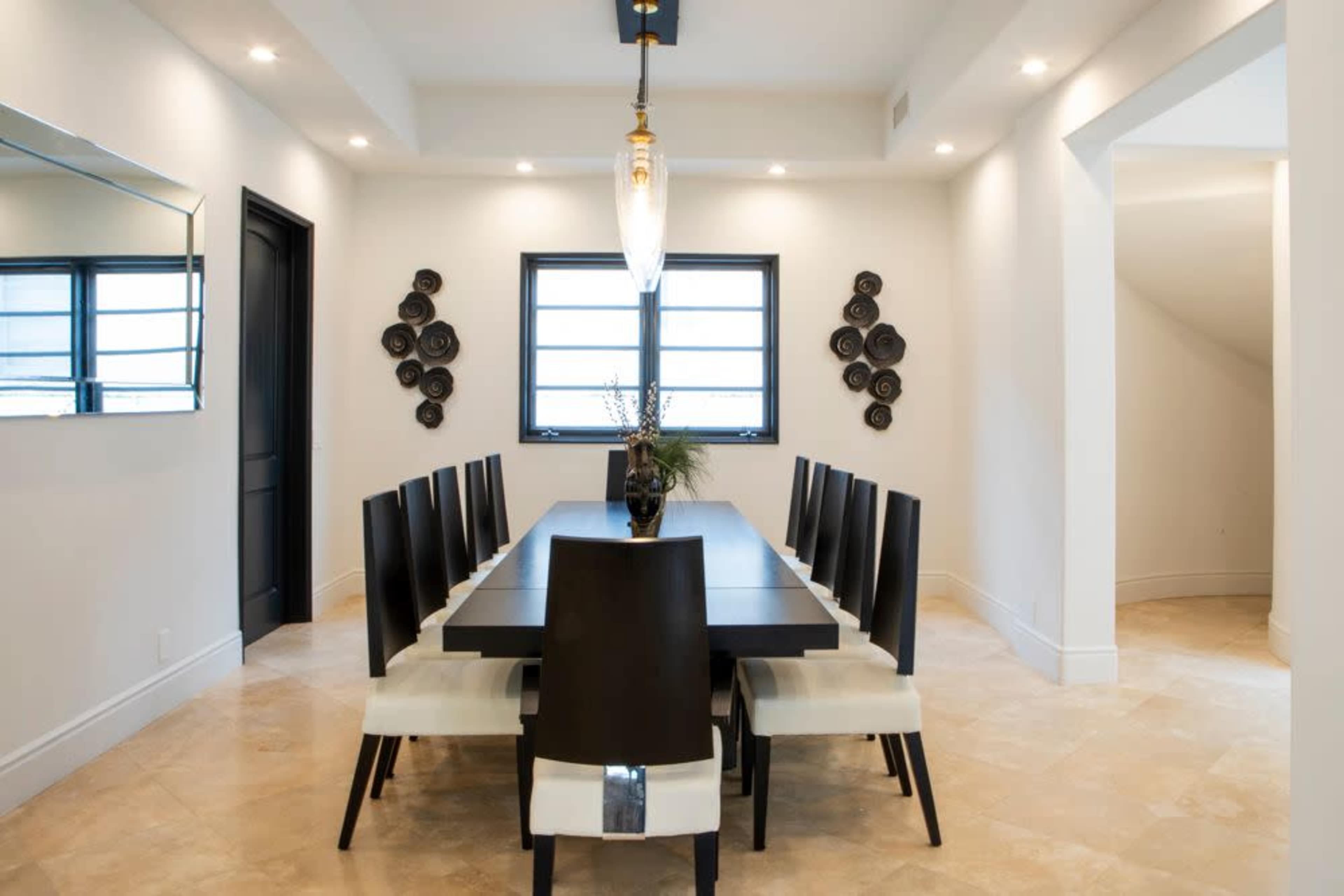 A modern dining room features a long black table surrounded by ten black and white chairs, with large windows and decorative plates on the walls.
