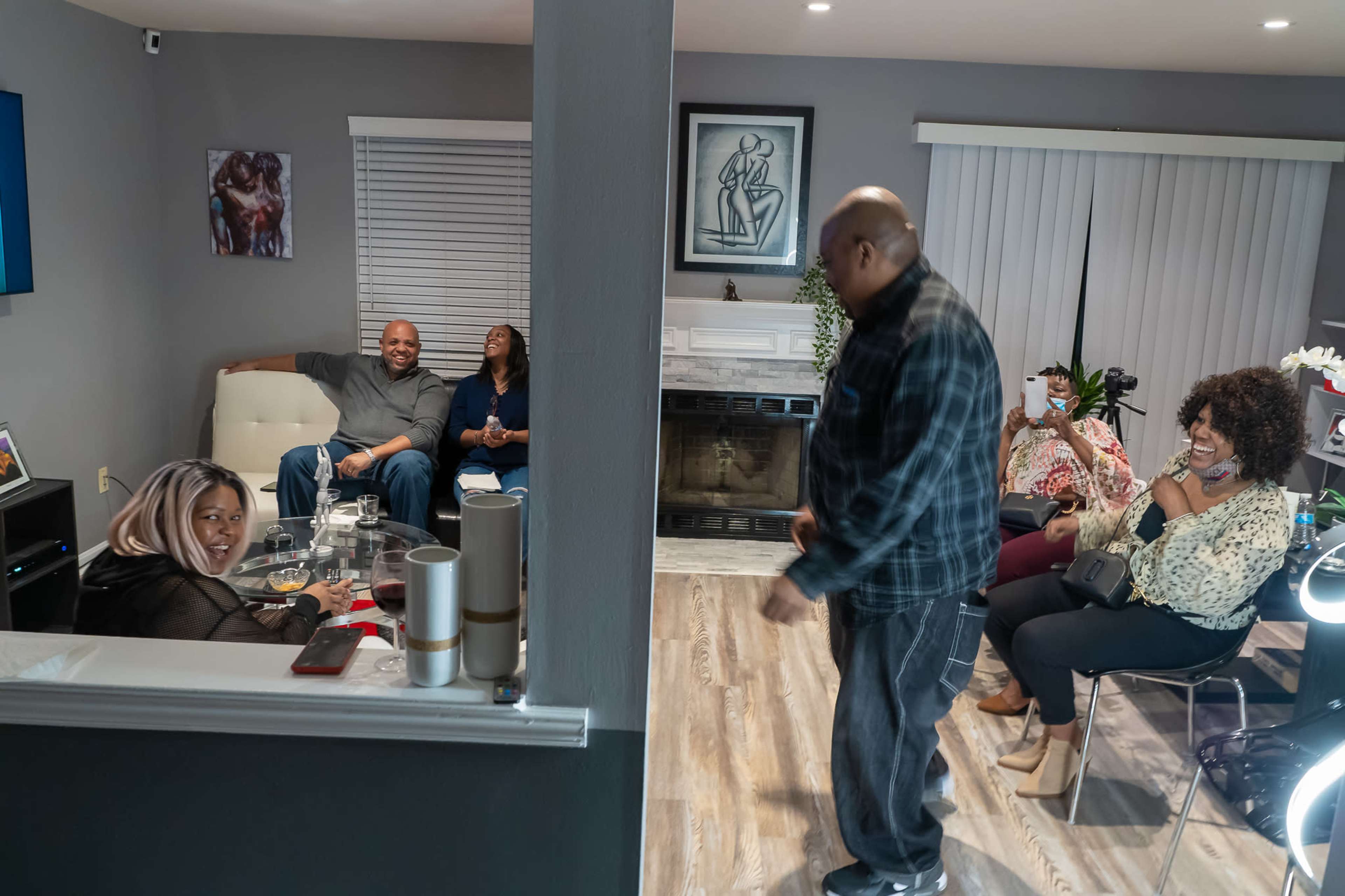 A group of people socializes in a living room, with some sitting on a couch and others standing, while snacks and drinks are visible on a table.