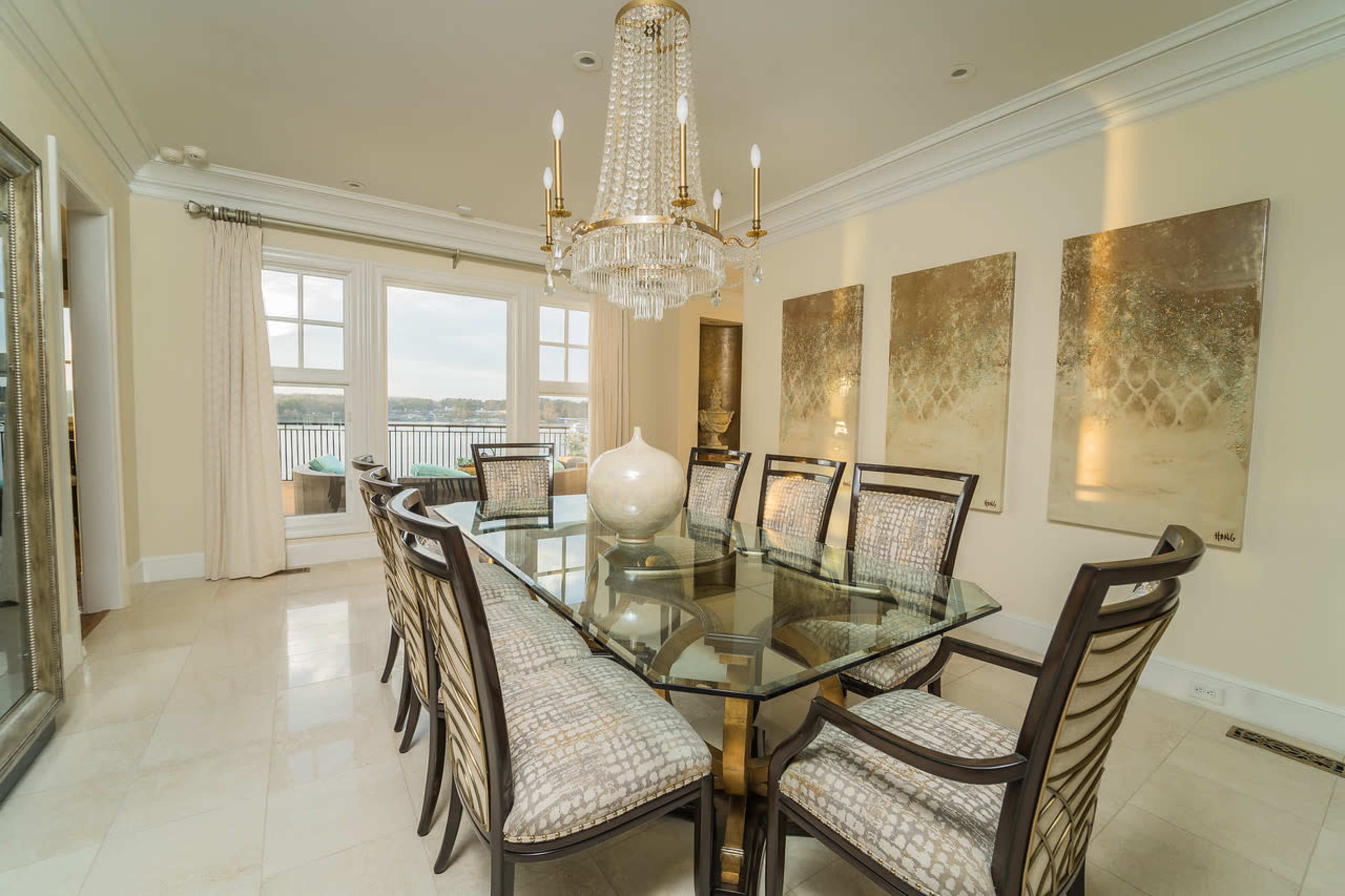 A spacious dining room features a large glass table surrounded by elegant chairs, with a chandelier overhead and decorative artworks on the walls.