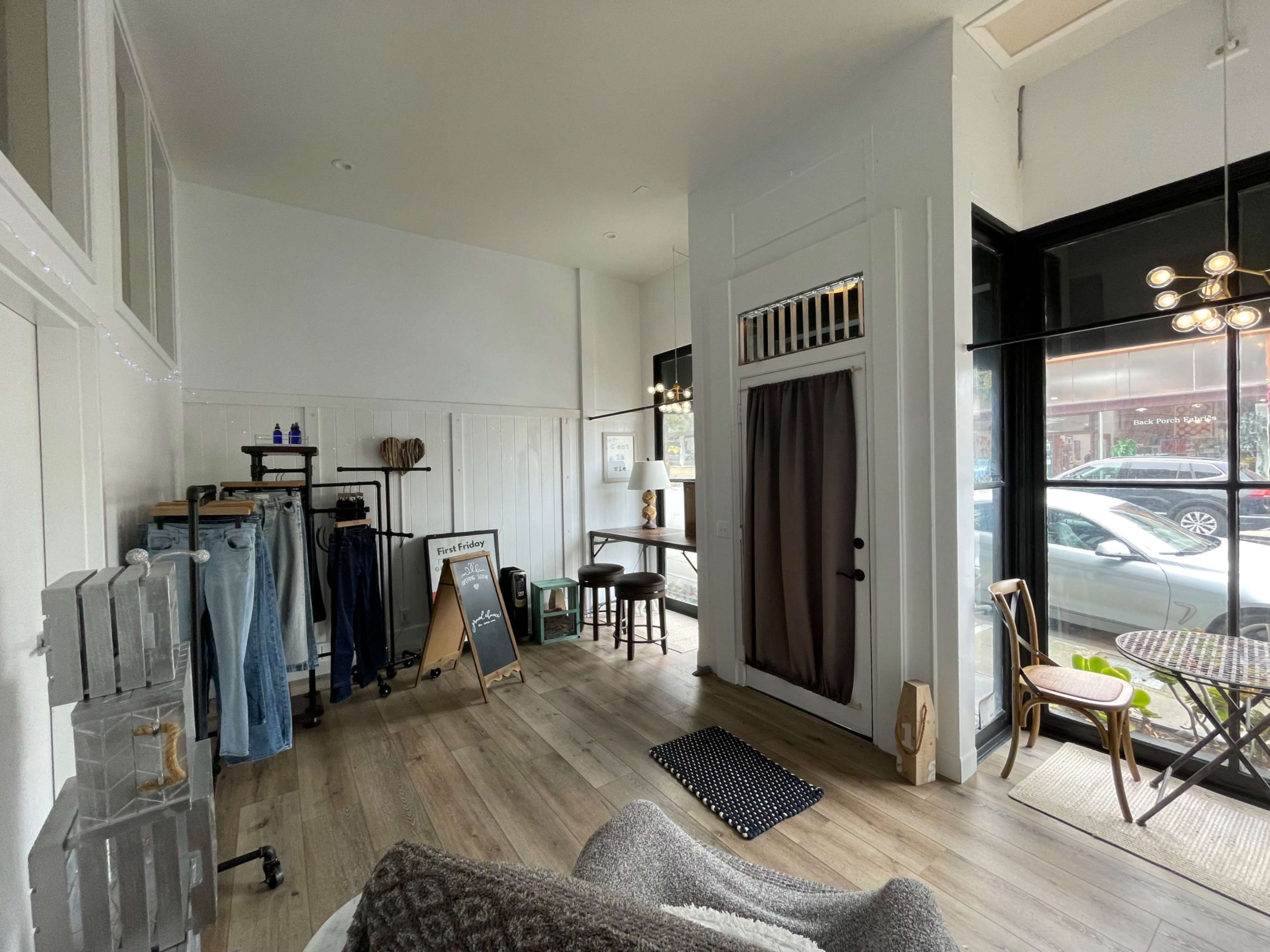 A brightly lit boutique interior with clothing racks, a small wooden table, and large windows facing the street.