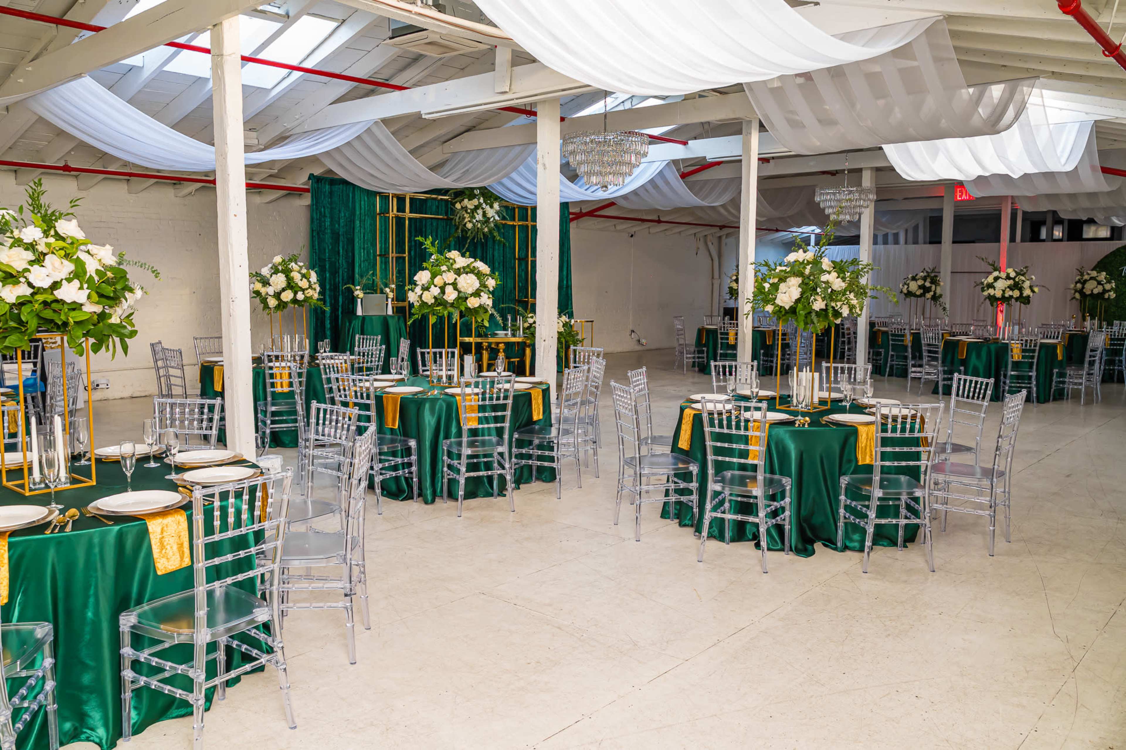 A spacious event venue set up with round tables covered in green tablecloths, adorned with floral centerpieces and elegant dining ware, under a canopy of draped fabric.