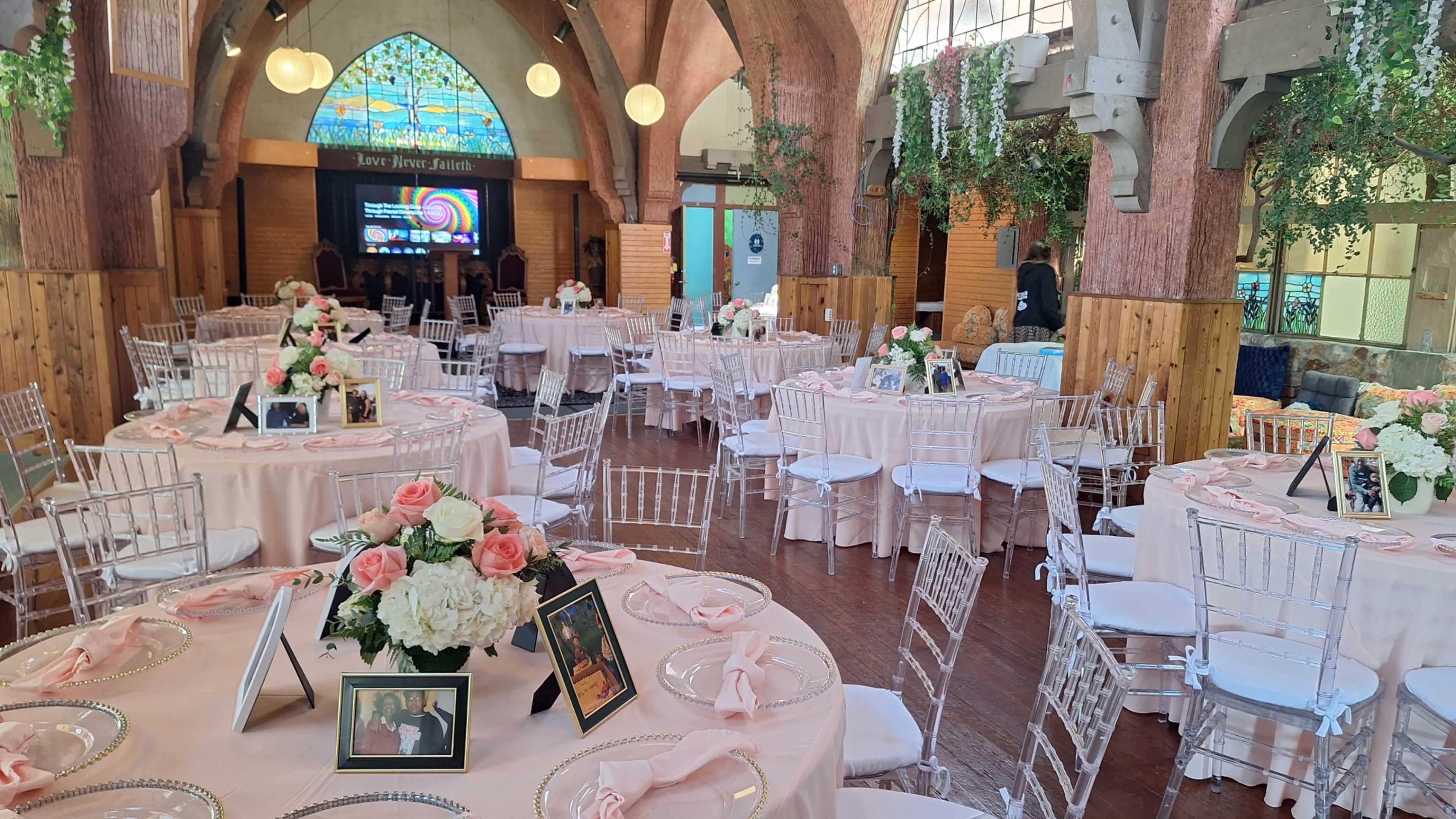 The image shows a banquet hall set up for an event, featuring round tables dressed with pink tablecloths, floral centerpieces, and framed photos, amid an elegant decor with arches and stained glass windows.