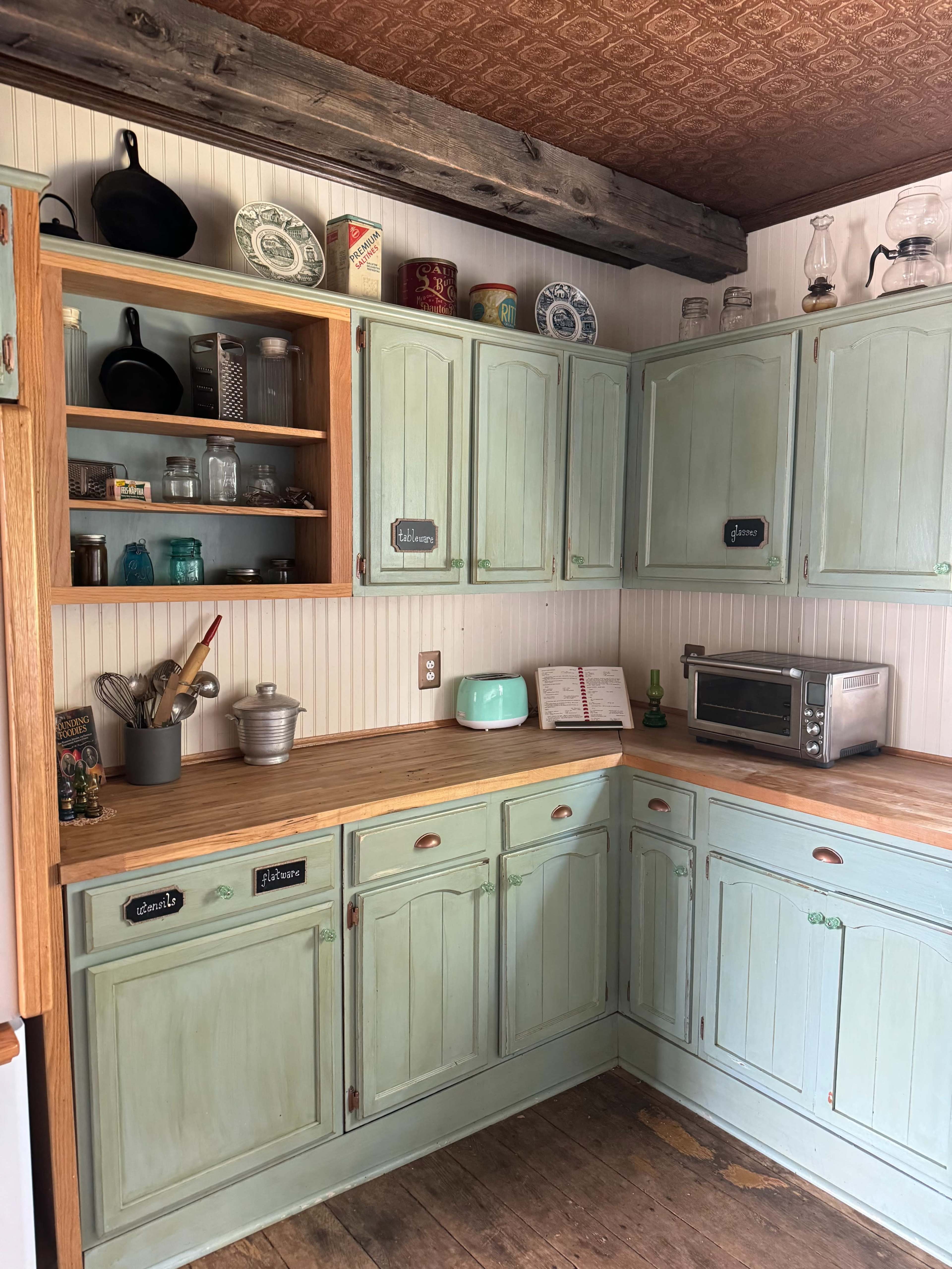 A rustic kitchen with light green cabinets, wooden countertops, and various jars and utensils displayed on shelves.