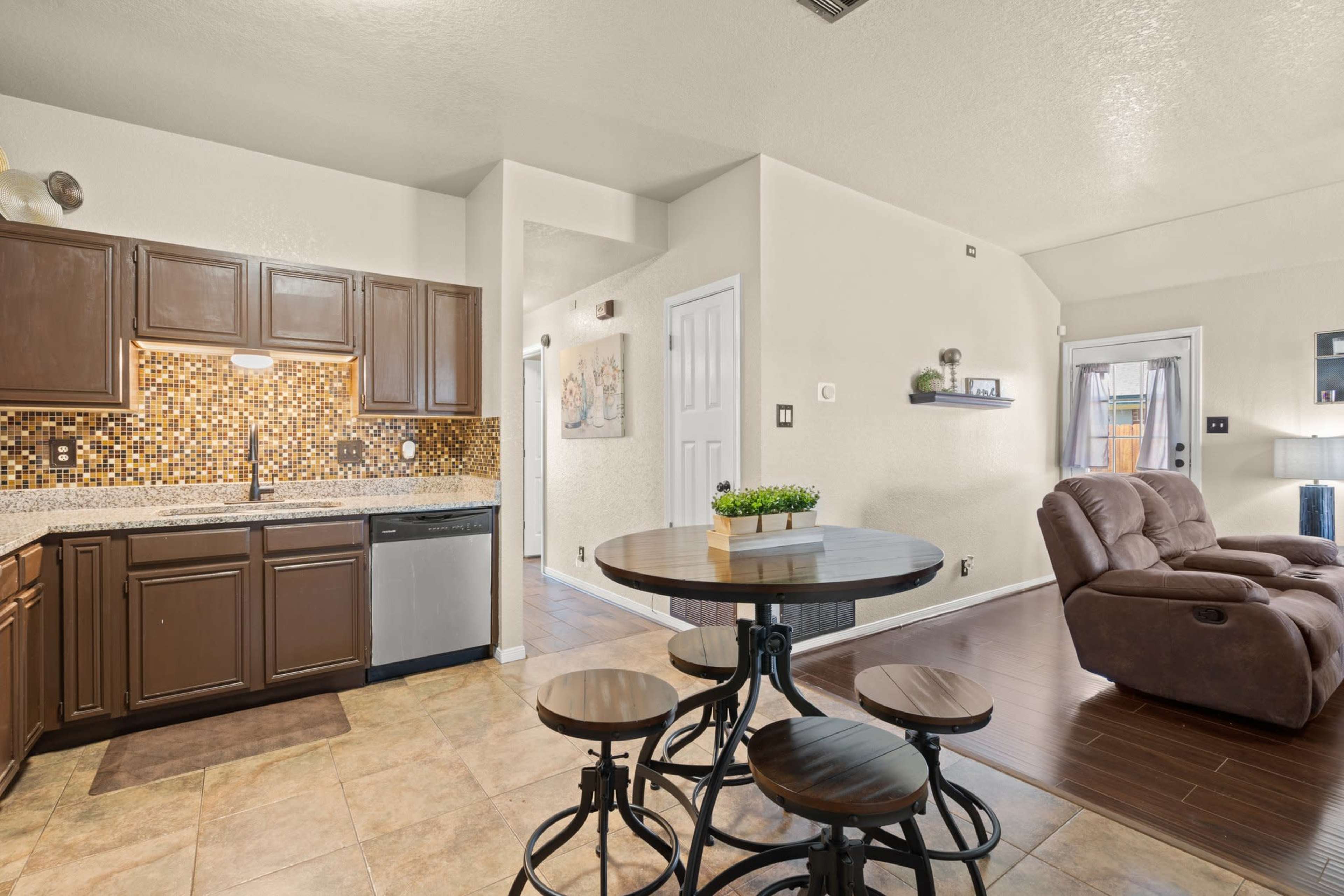 A compact kitchen area with dark wooden cabinets, a tiled backsplash, and a round dining table with several stools, adjacent to a spacious living area featuring a recliner chair.