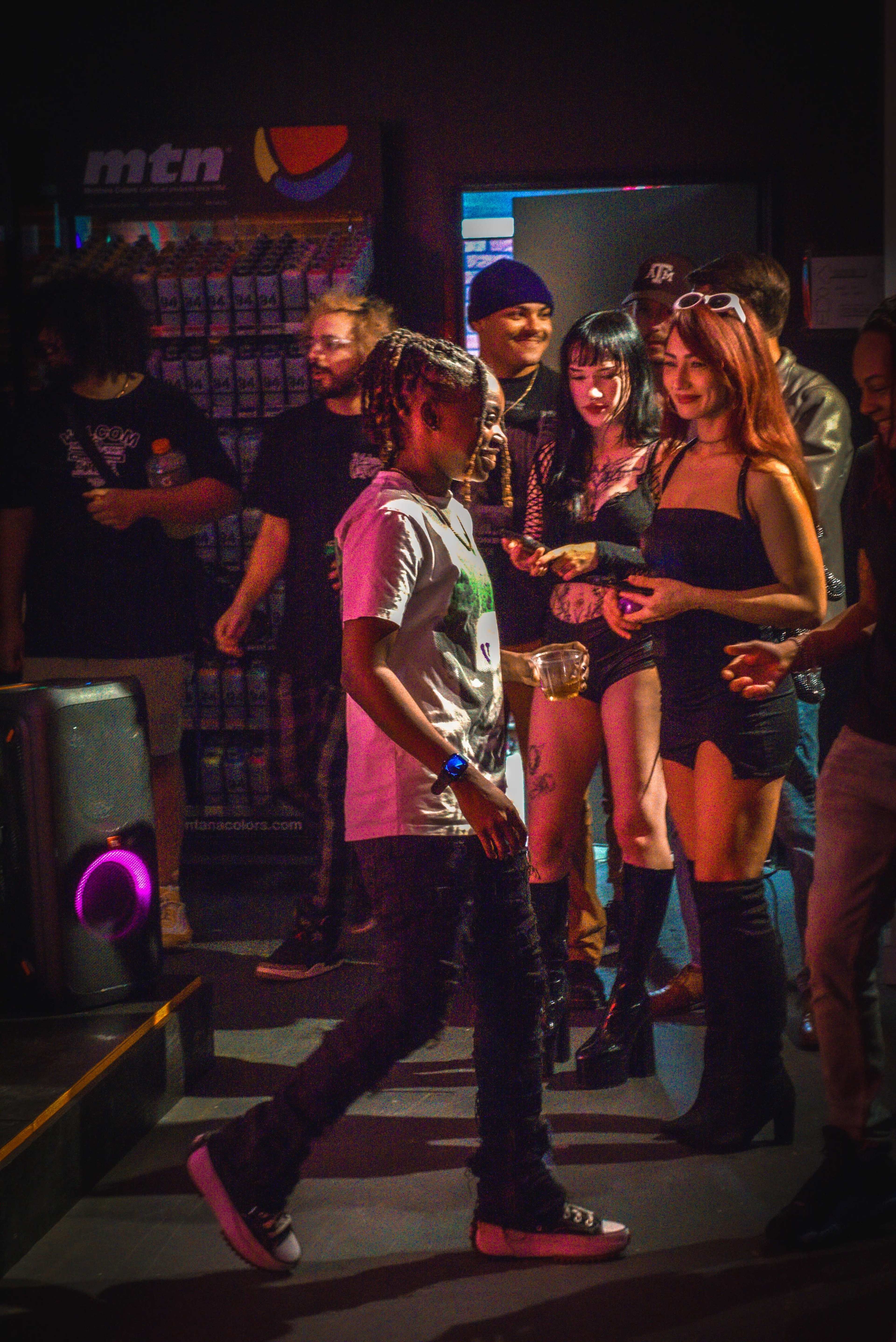 A group of people socializes in a dimly lit venue, with a mix of individuals dressed in various styles, including one person in a graphic t-shirt and another in a black outfit.