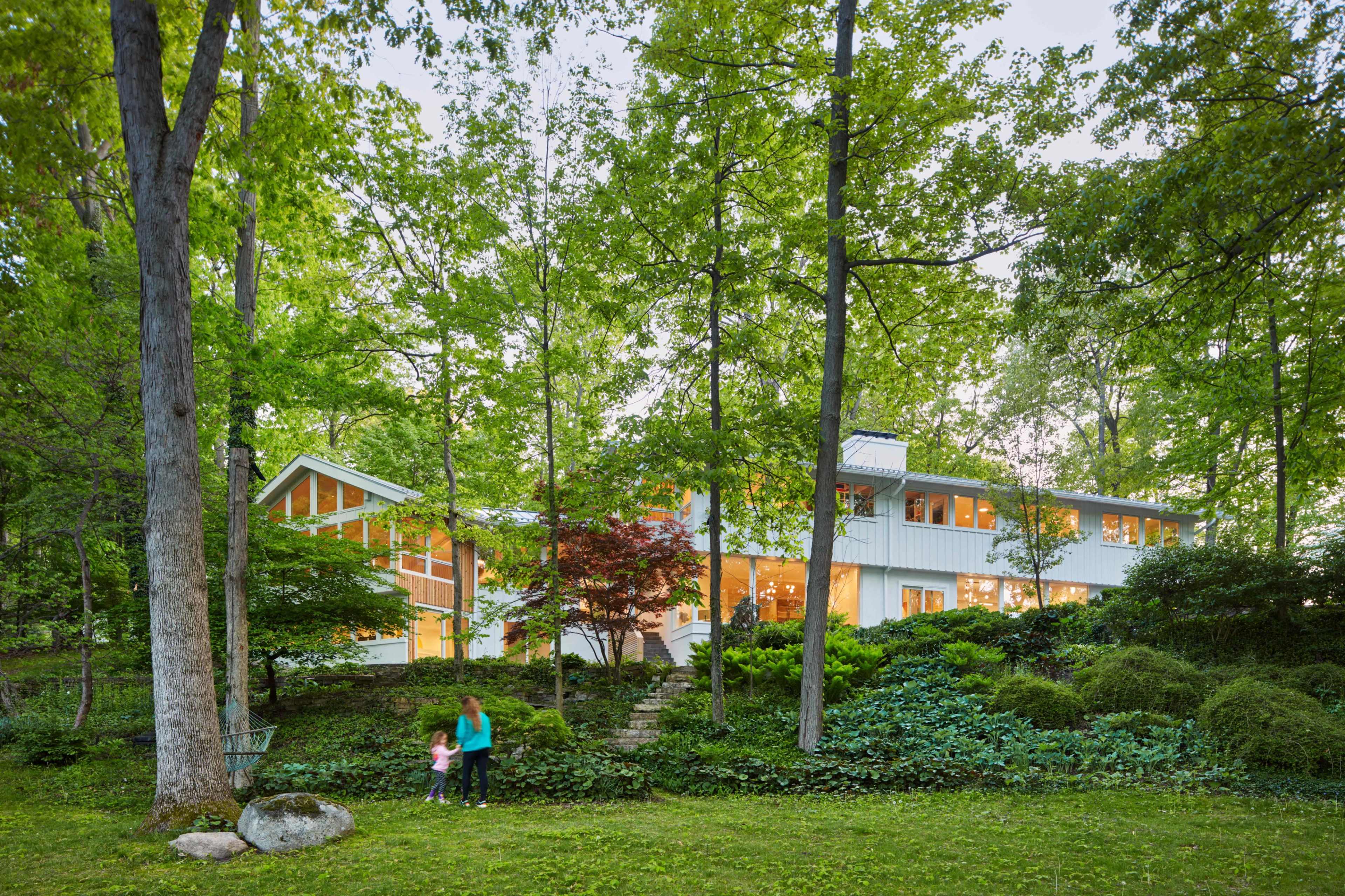 A modern house is nestled among tall trees and greenery, with people walking in the front yard.
