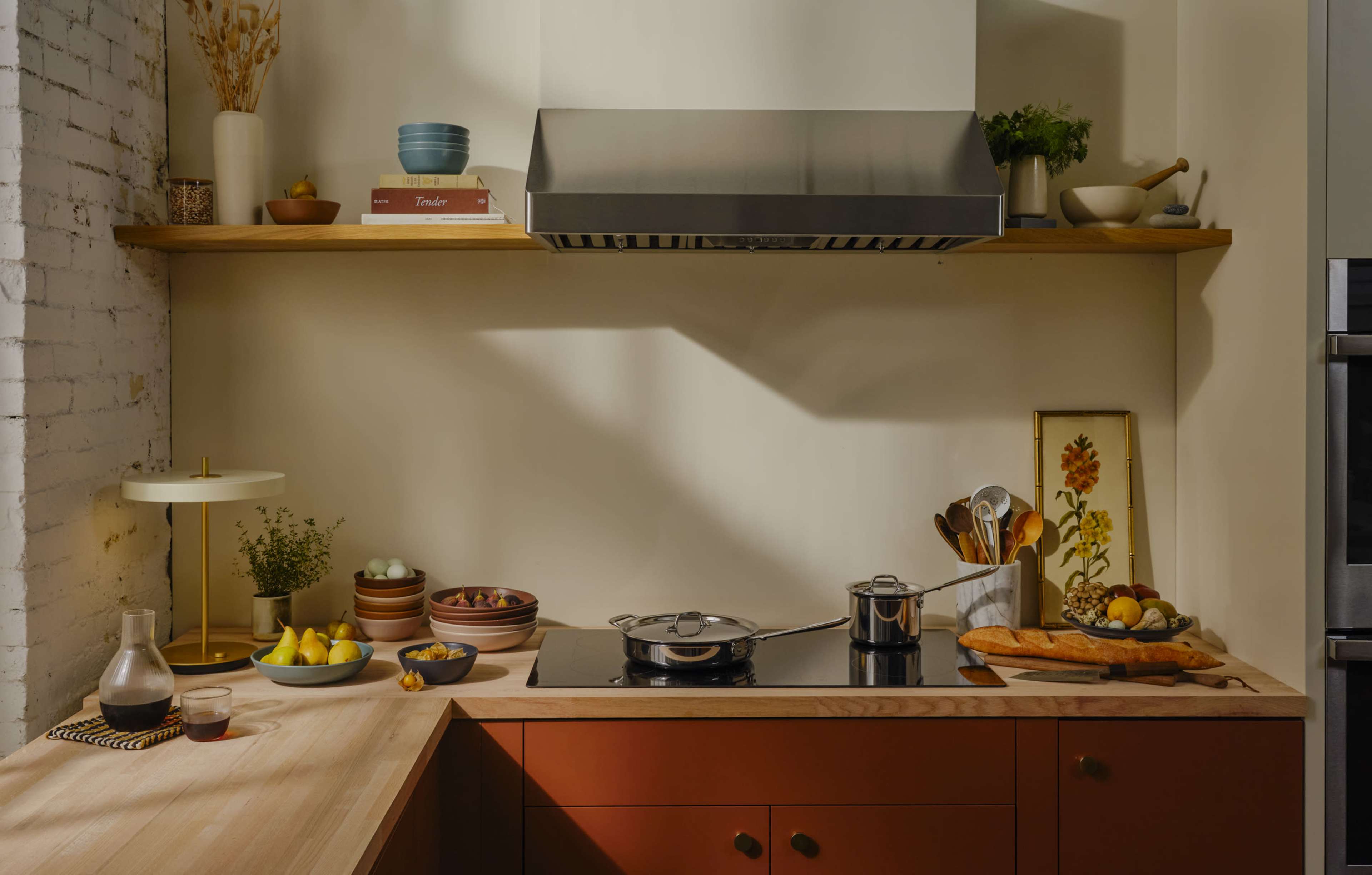 A modern kitchen features a wooden countertop, a stovetop with a pot, various bowls of fruits, and organized cooking utensils on a shelf.