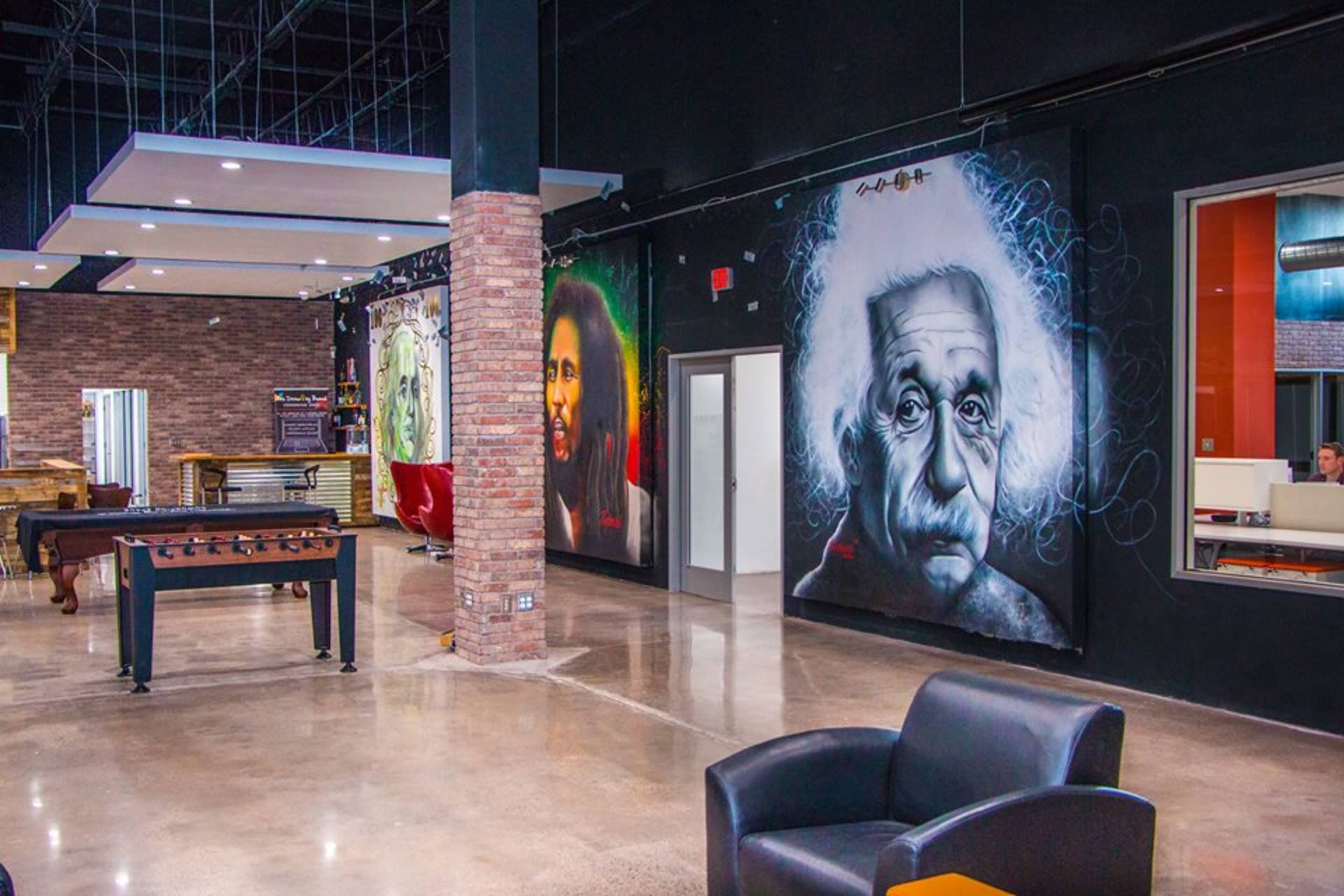 A modern interior space featuring large murals of iconic figures, including Albert Einstein and Bob Marley, with a foosball table and seating areas visible.