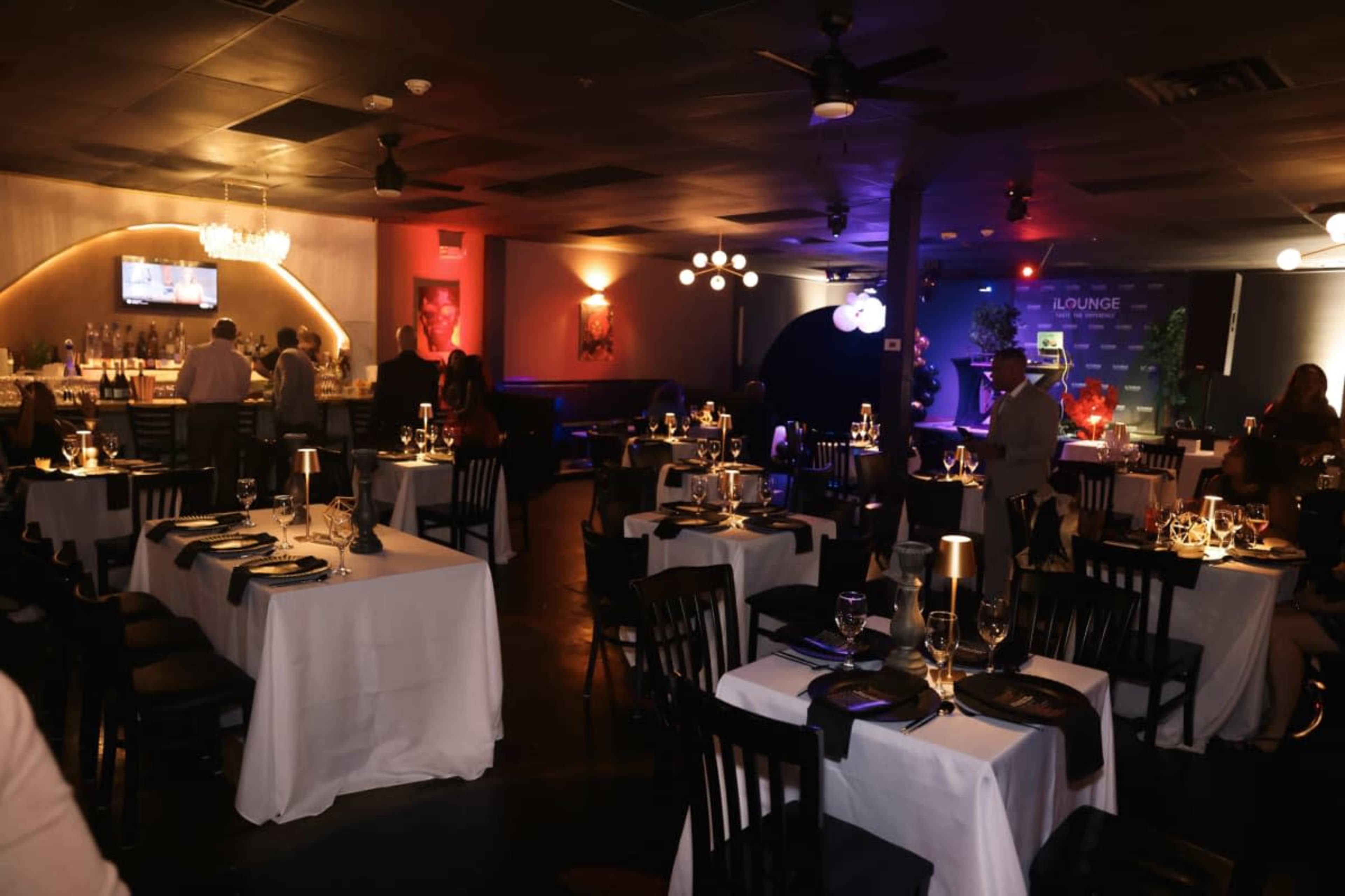The image shows a dimly lit restaurant interior with neatly set tables, soft lighting, and a bar area in the background.