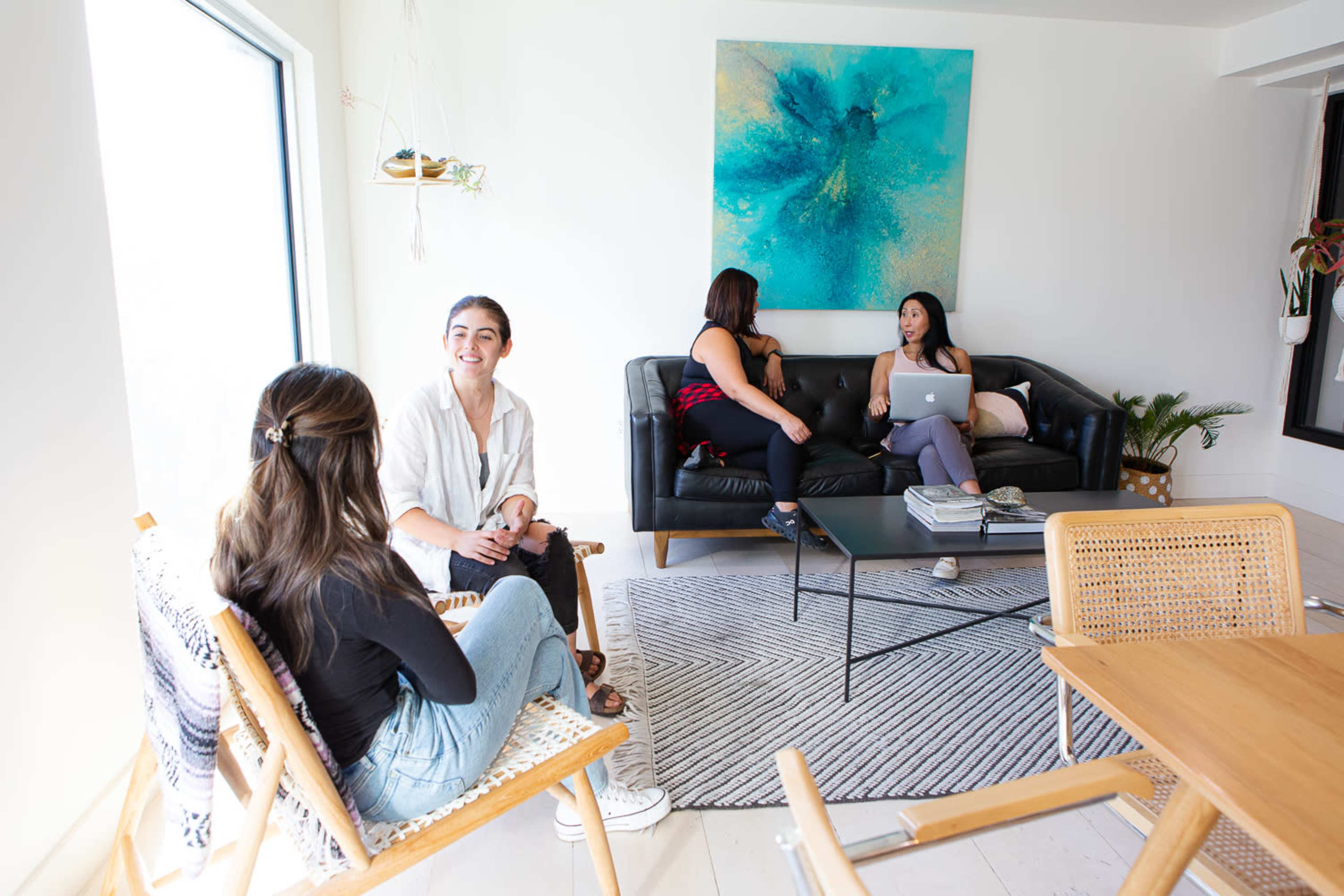 Four women are gathered in a bright, modern living space, with two sitting on chairs and two on a sofa, engaged in conversation.