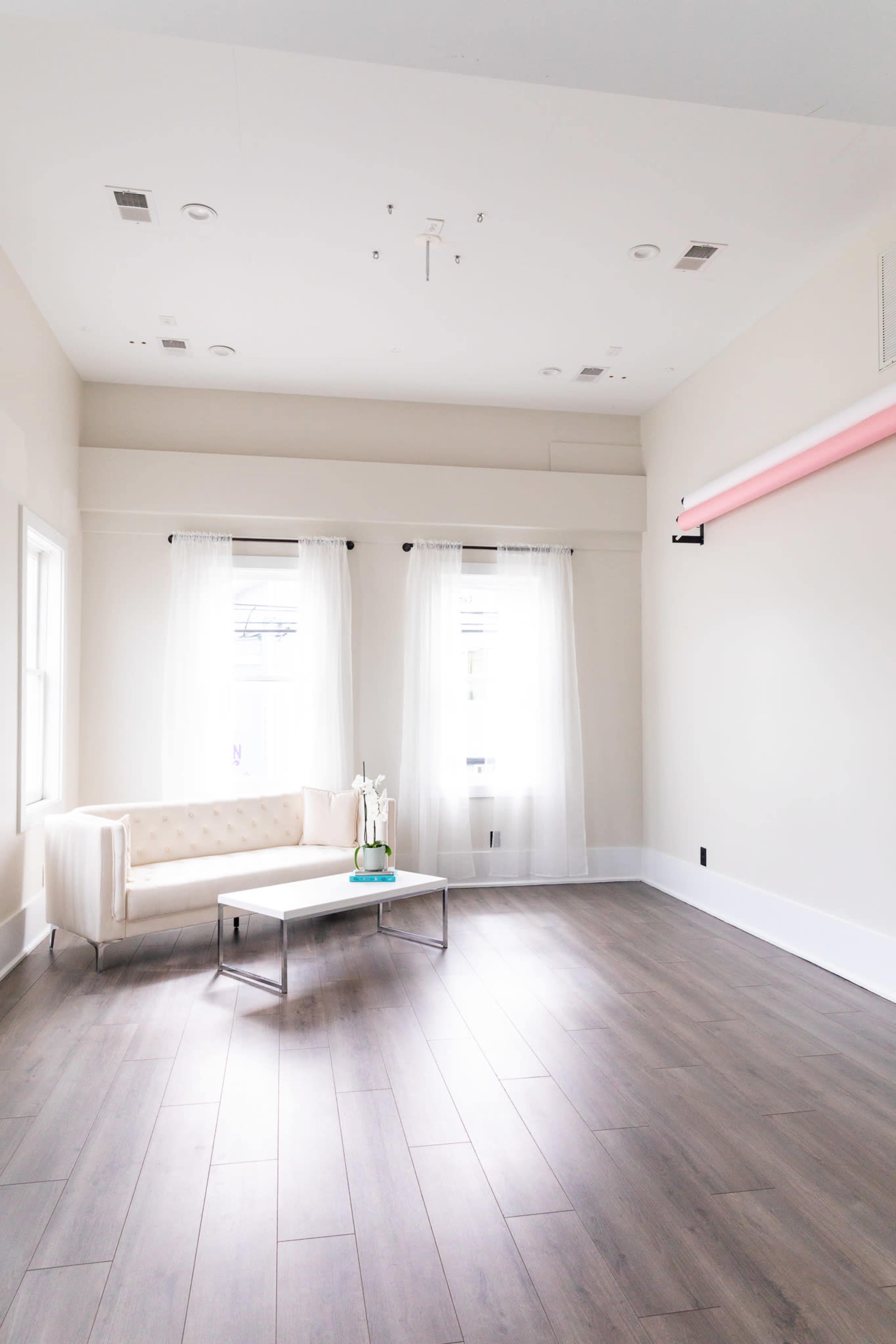 A spacious room with a light-colored wall, two large windows with sheer curtains, a beige sofa, and a glass coffee table on a wooden floor.