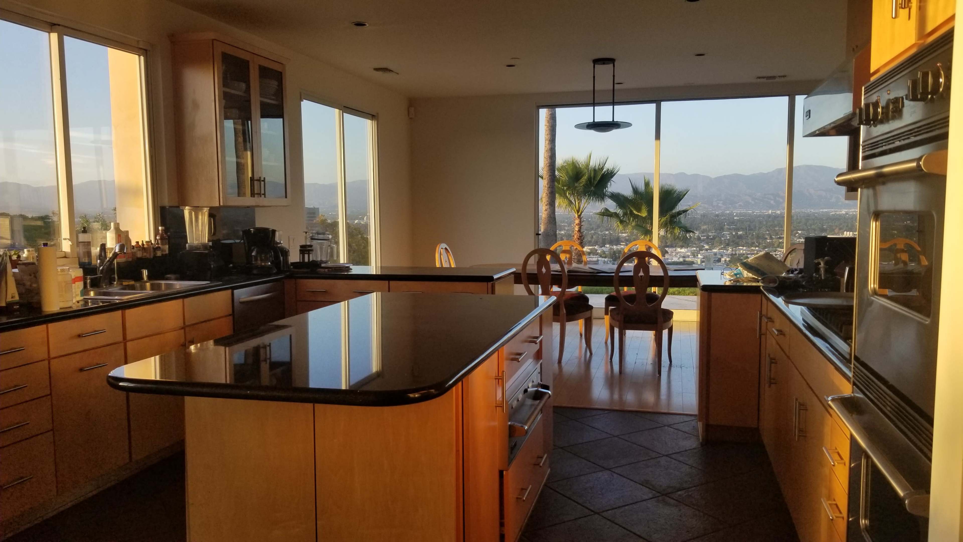 A modern kitchen with wood cabinetry and a large island, featuring a view of a patio and mountains through large windows.