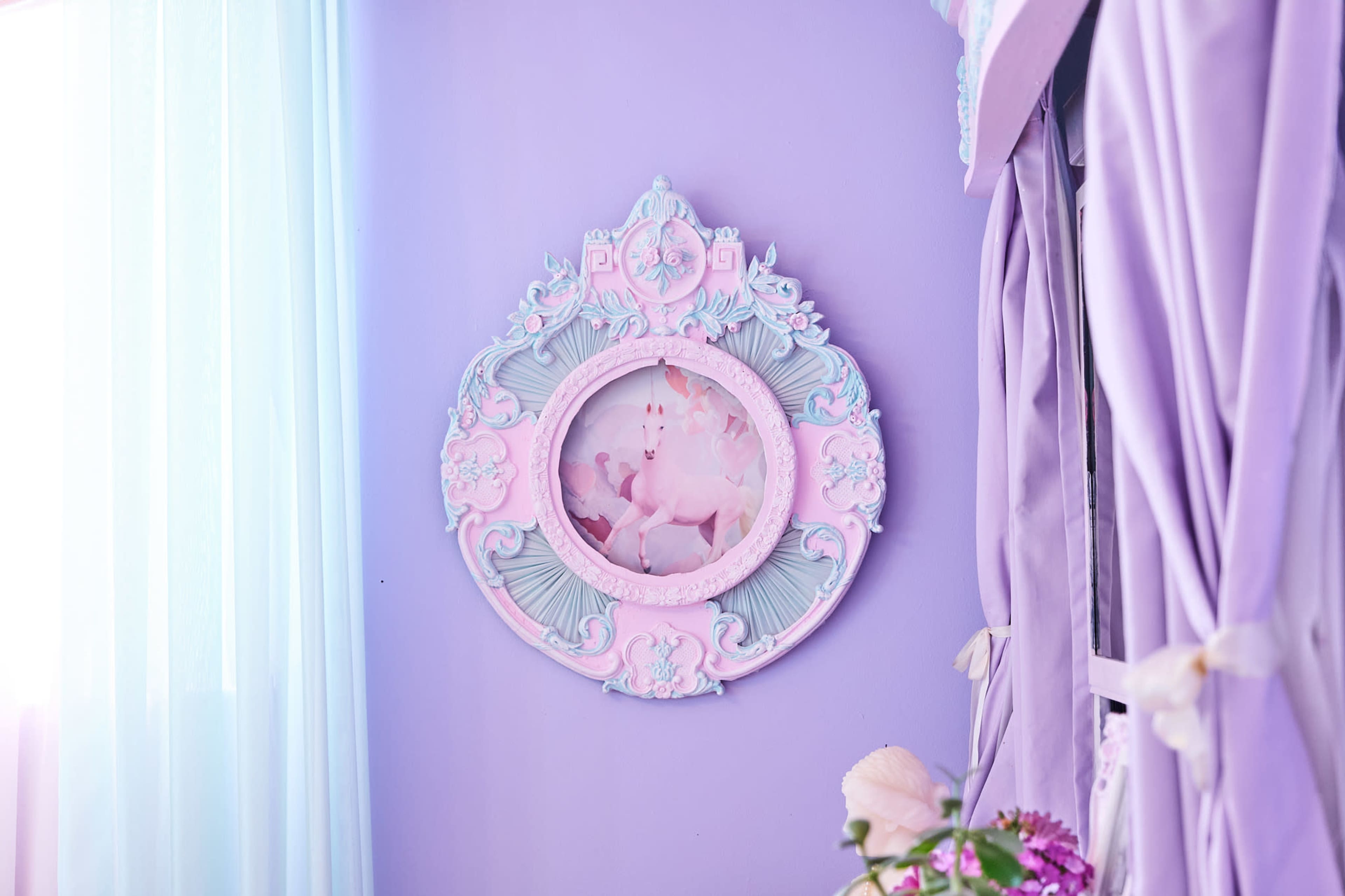 A vintage-style mirror with pastel framing hangs on a lavender wall, surrounded by sheer curtains.