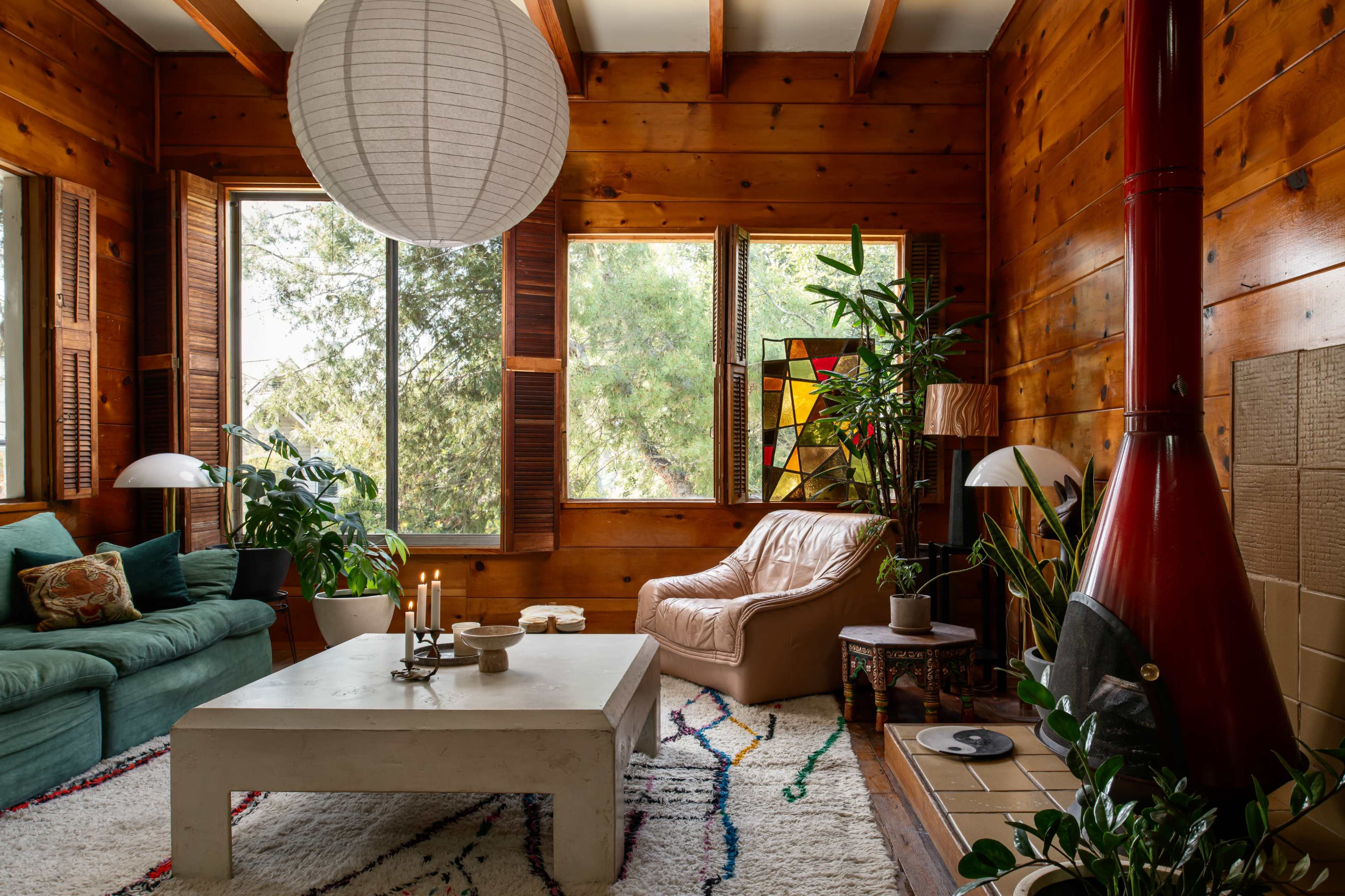 A wooden-paneled living room with a green sofa, a white coffee table, a large paper lantern, and various plants near a colorful stained glass window.