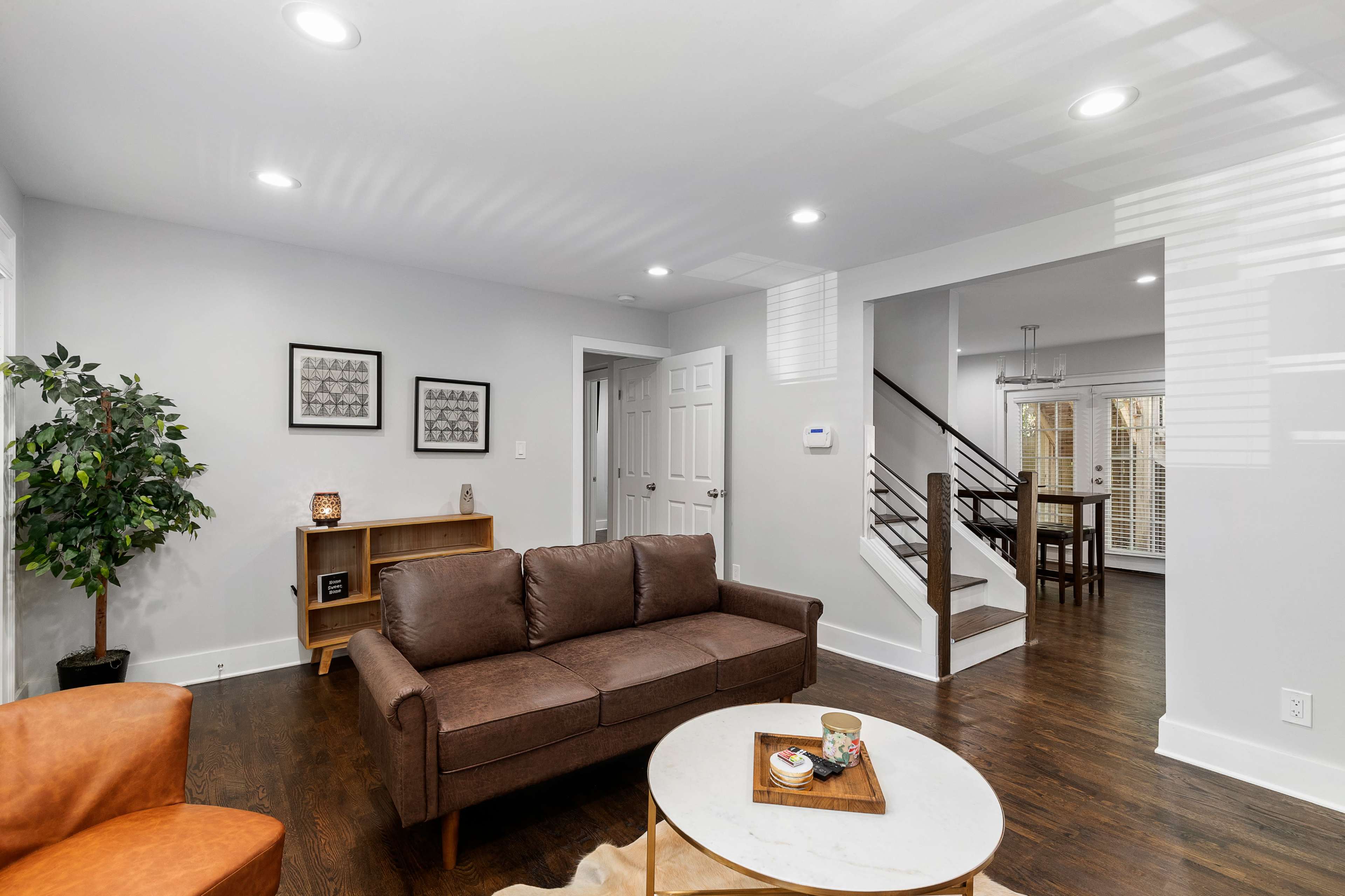 A modern living room with a brown sofa, a round coffee table, and a bookshelf, featuring a staircase and a dining area in the background.