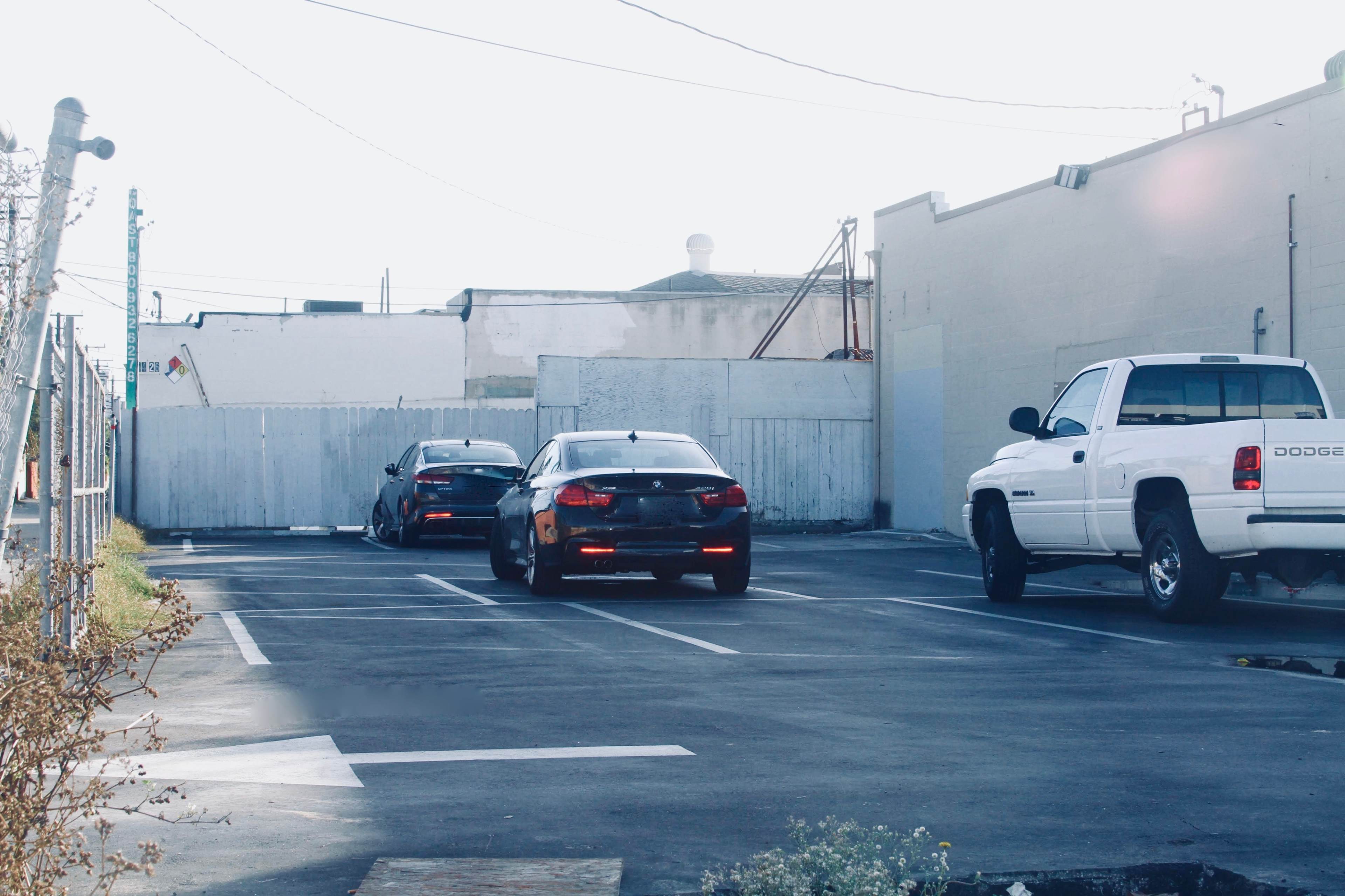 Two cars are parked in a lot with a white truck in the background near a fence and a building.