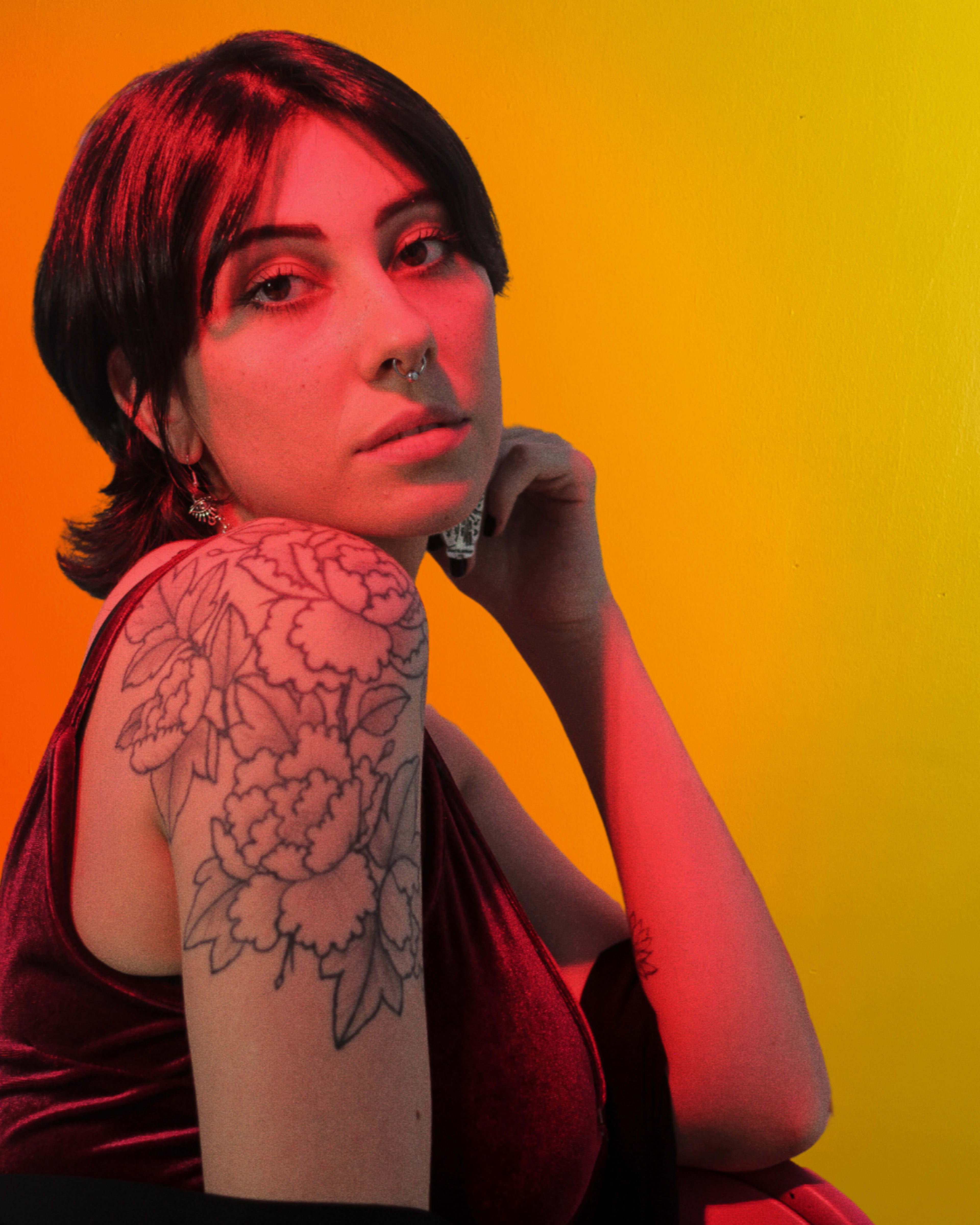 A woman with a floral tattoo on her shoulder poses against a gradient yellow and orange background.