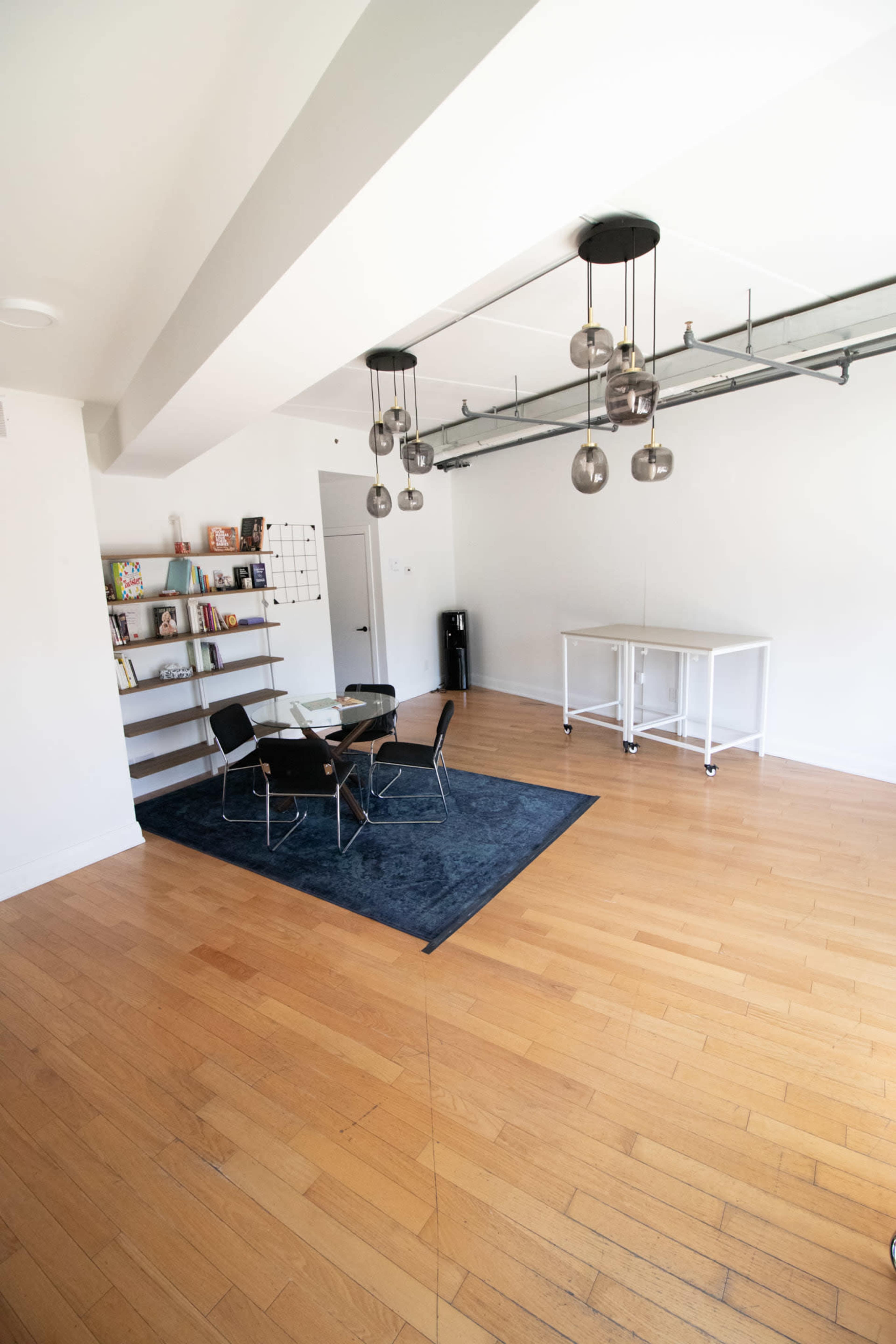 The image shows a modern office space featuring a meeting area with a glass table and black chairs on a dark rug, a bookshelf with various items, and overhead pendant lighting.