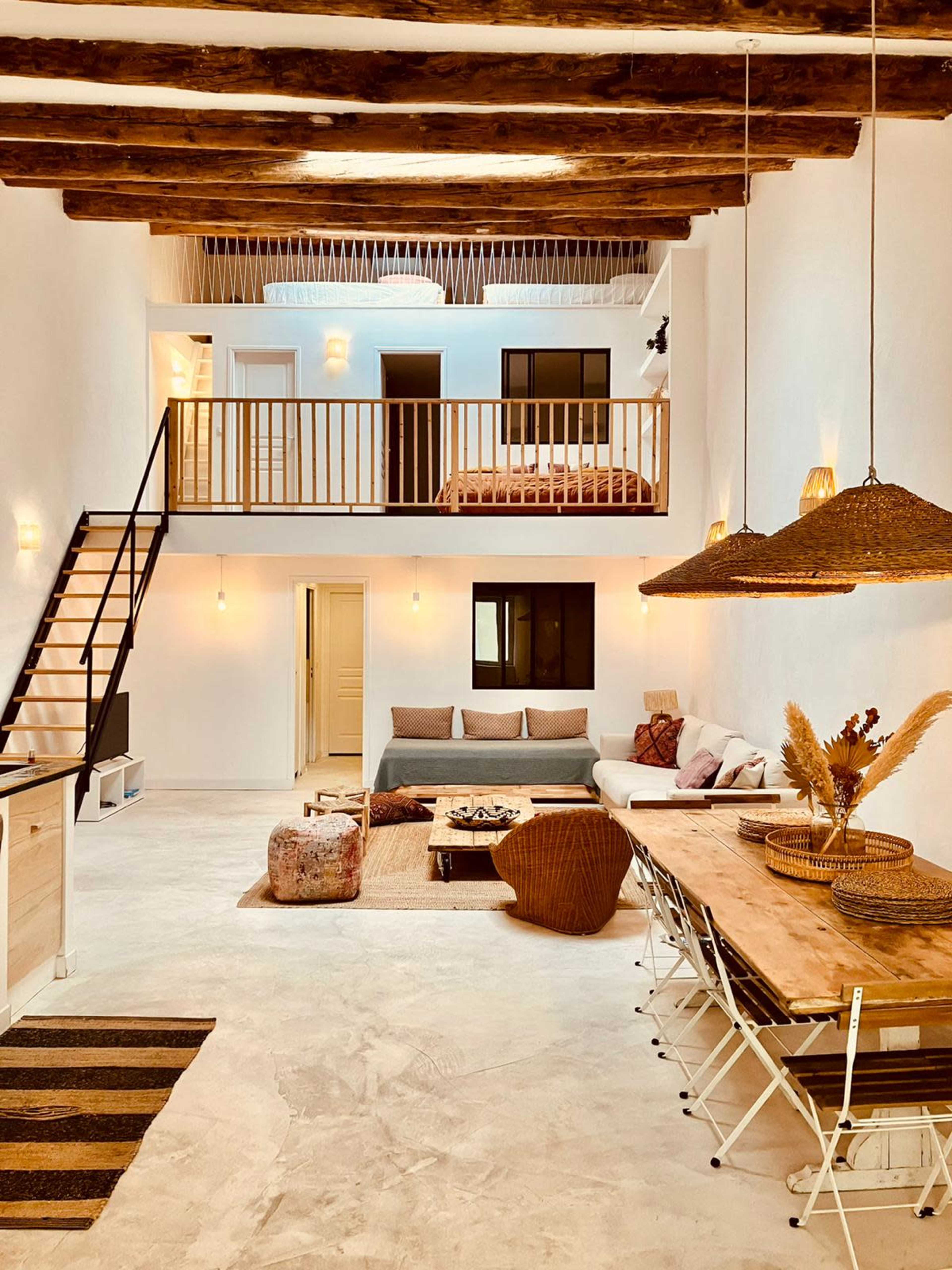 The image shows a spacious, modern living area with high ceilings, wooden beams, a dining table, a sectional sofa, and a lofted bedroom visible in the background.