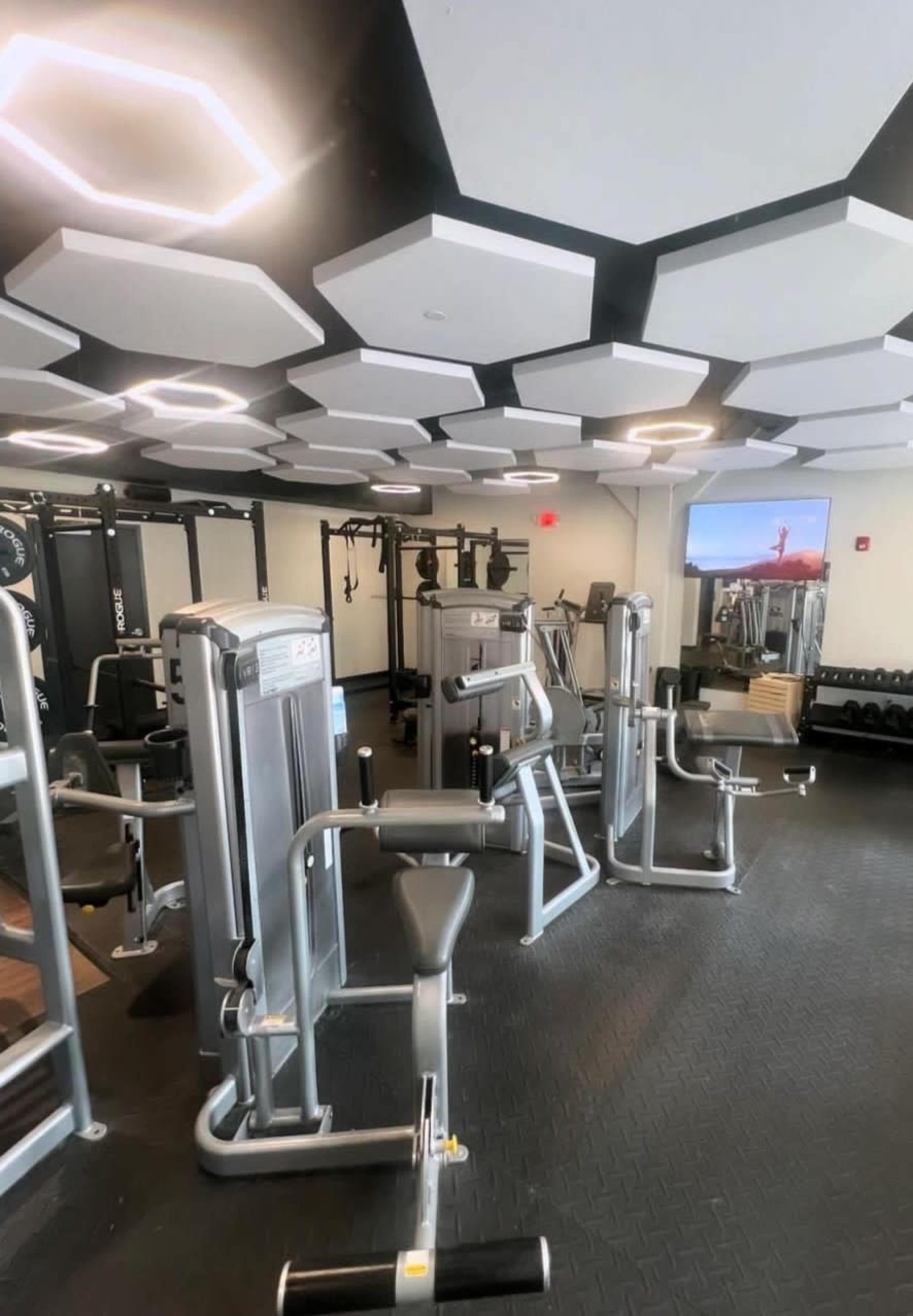 The image shows a spacious gym equipped with various exercise machines and a large screen displaying a workout video.