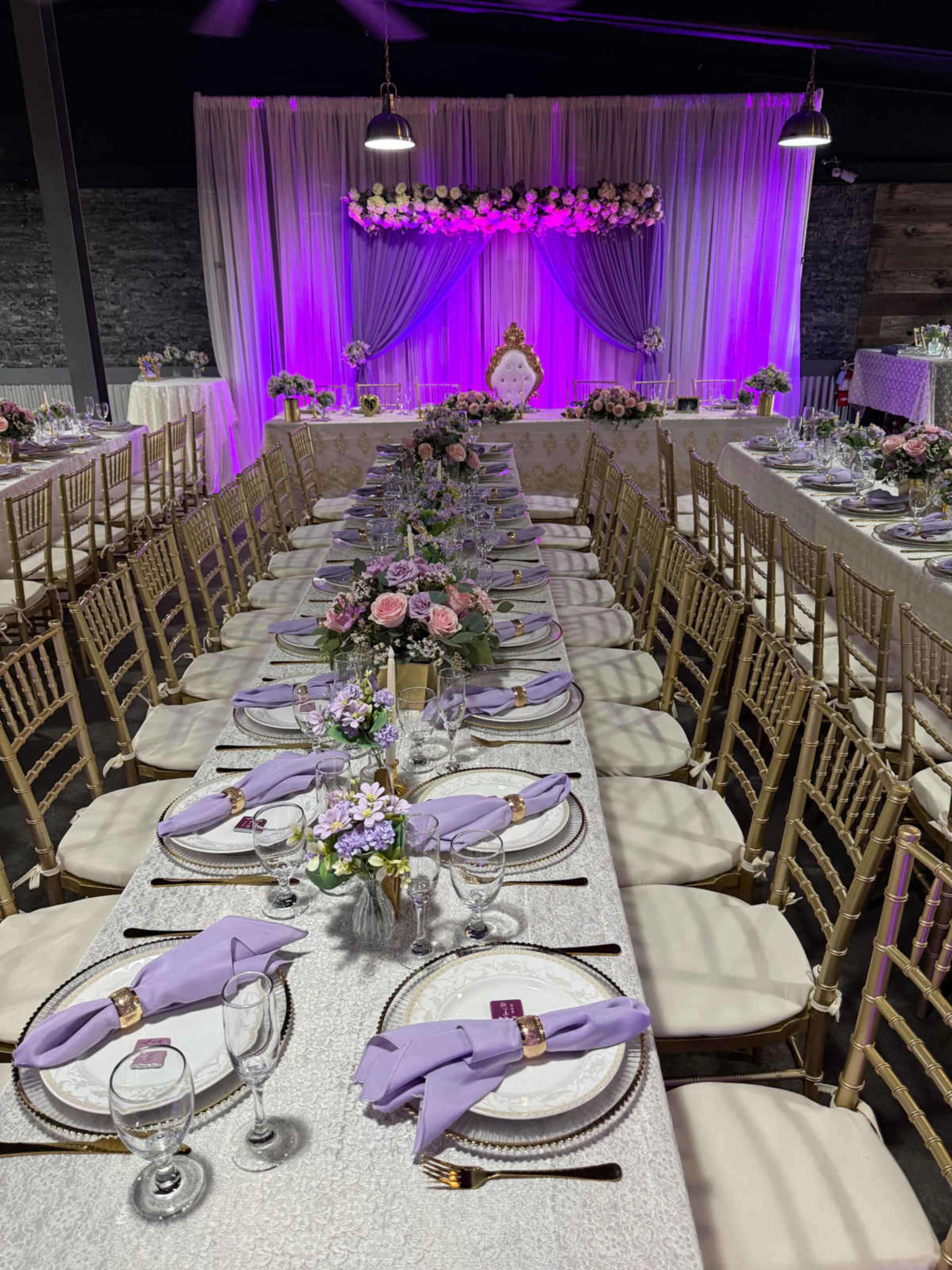 The image shows a banquet setup with long tables adorned with lavender napkins, floral centerpieces, and purple lighting in a decorated venue.